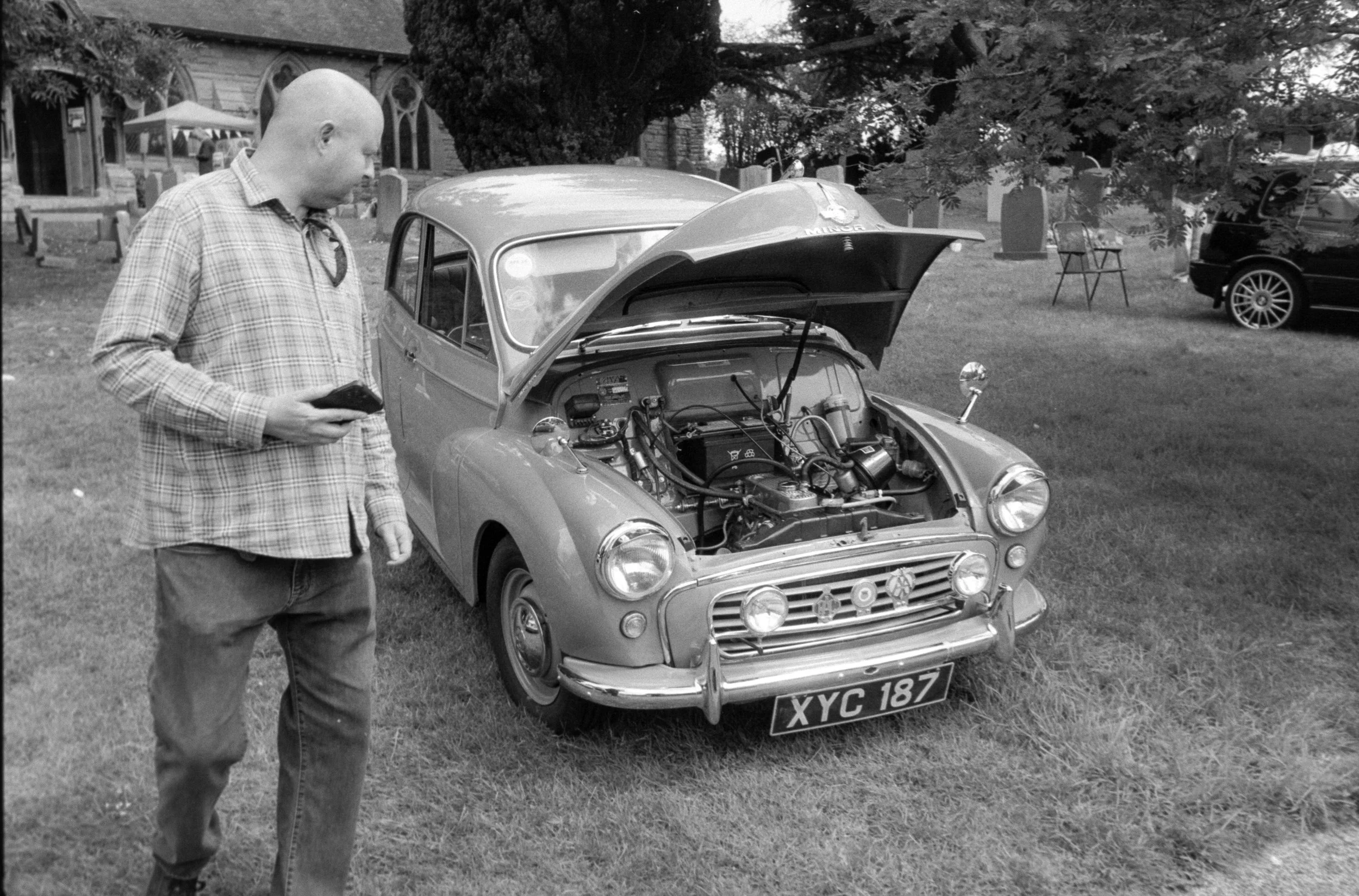 A man with a bald head, wearing a checkered shirt and jeans, stands next to a small vintage car with its hood open in a park or garden area, possibly inspecting or repairing it.