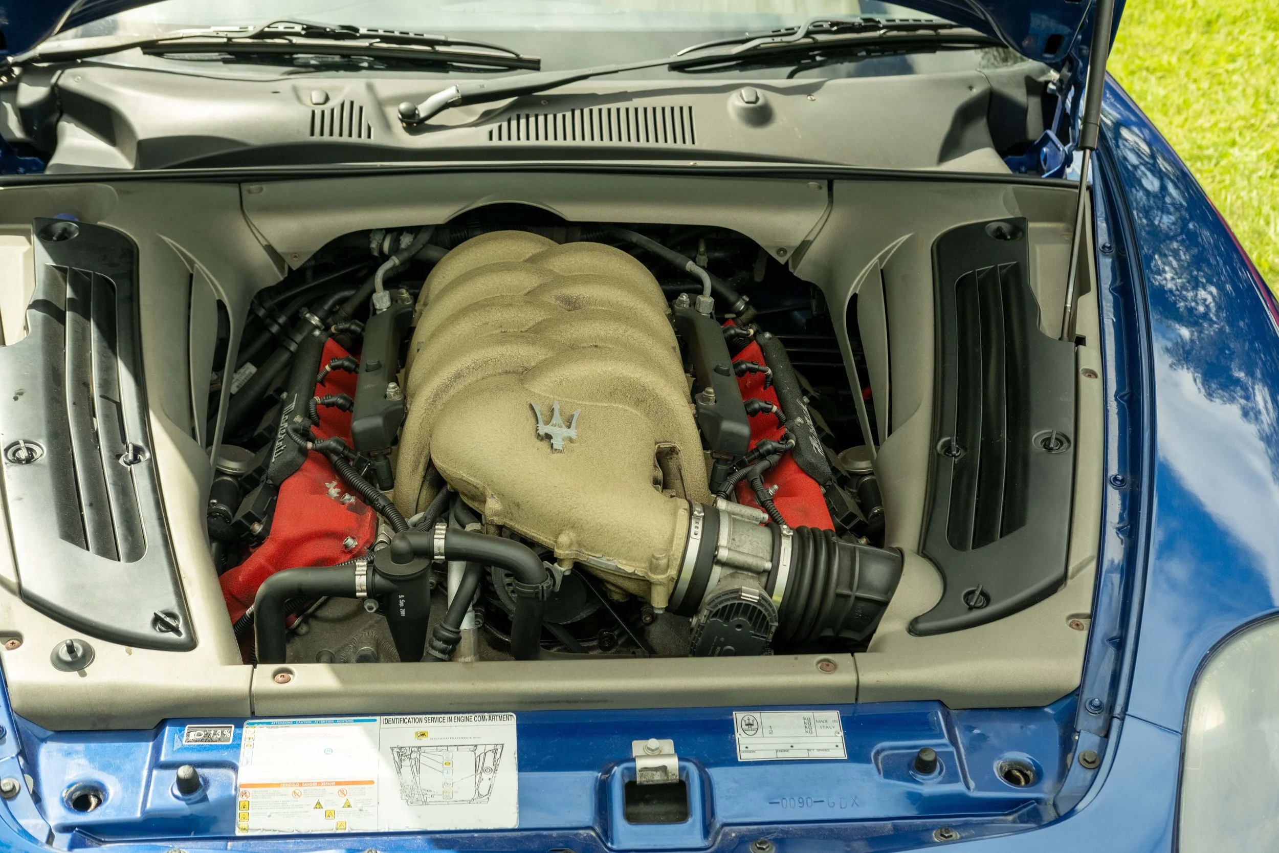 Car engine bay with visible engine components, hoses, and air intake system, under the open hood of a blue vehicle parked outdoors.