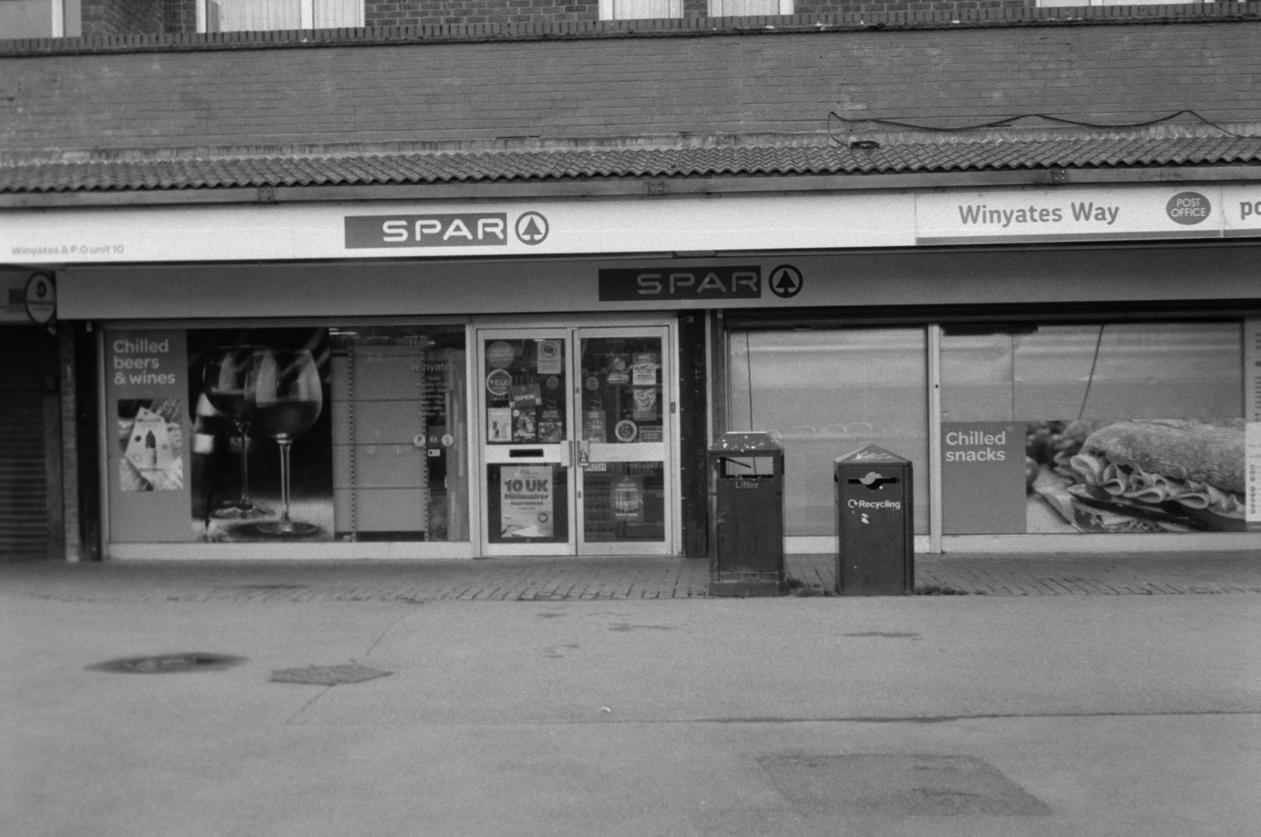 The exterior of a small grocery store with signs advertising chilled beers, wines, snack foods, and a recycling bin in front. The store has a glass door entrance and a large window showcasing various posters and advertisements.