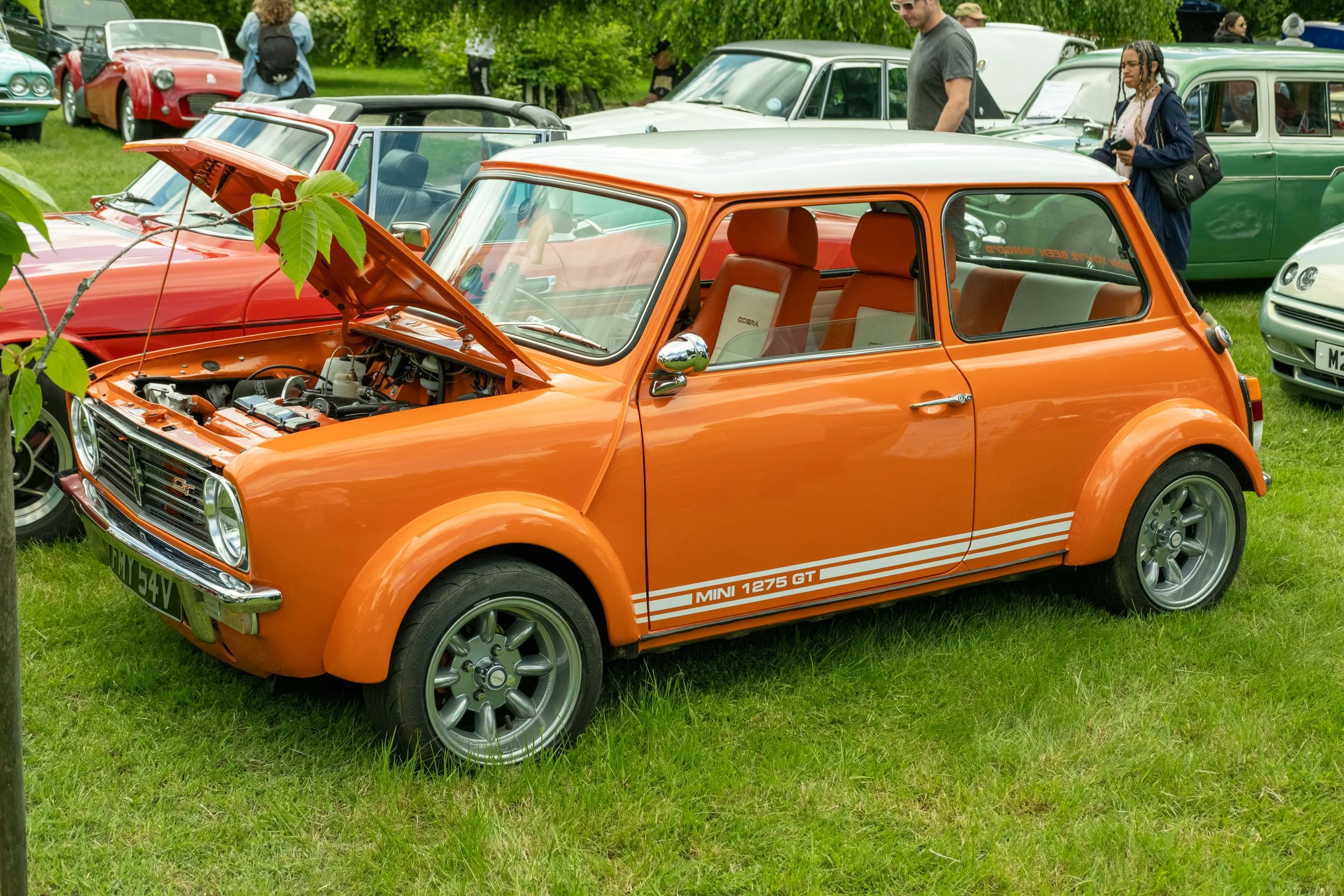 An orange Mini Cooper with its hood open, displayed at a classic car show on a grassy field, surrounded by other vintage cars and people.