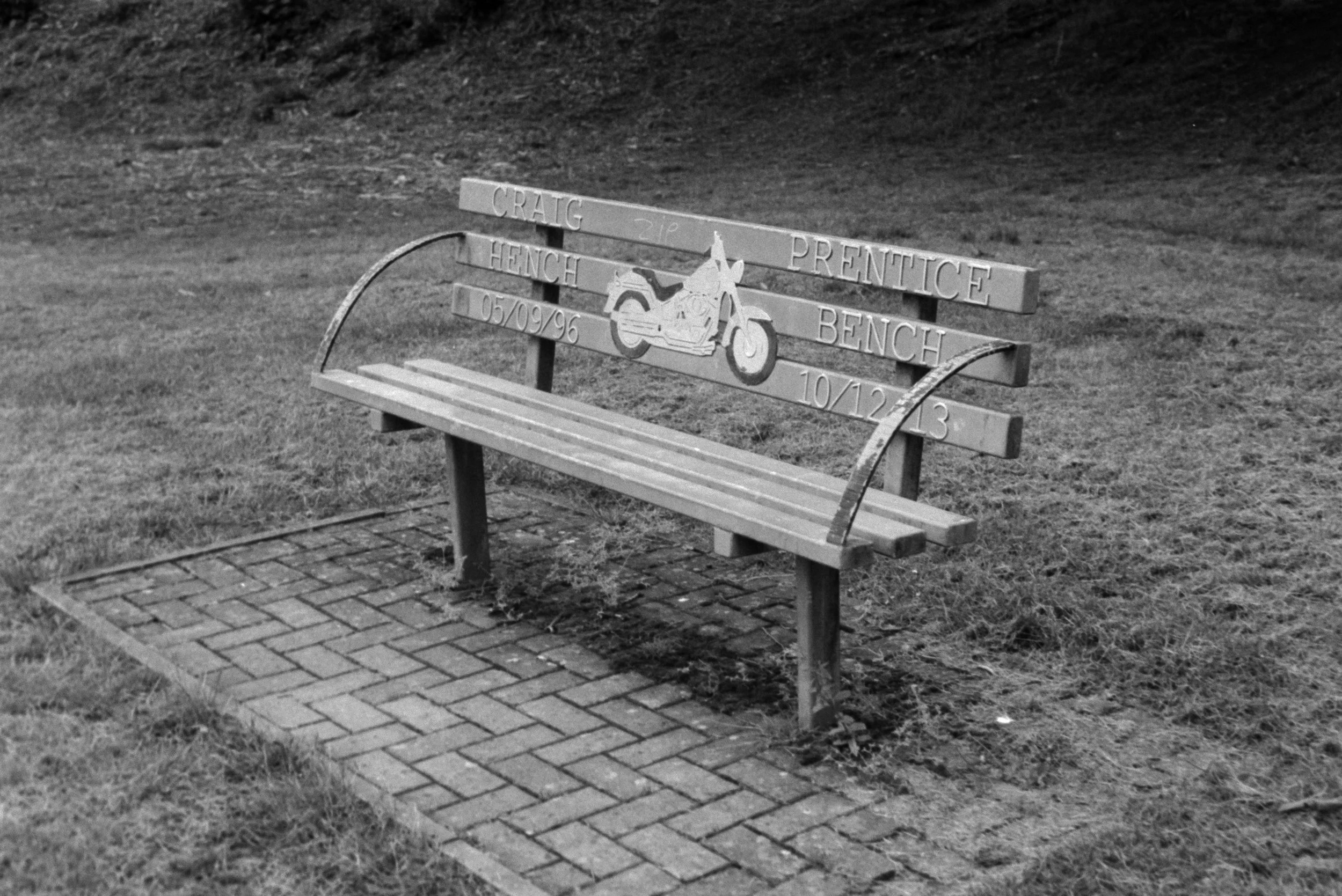 A park bench with a wooden seat and metal armrests and backrest, decorated with a motorcycle graphic and engraved with names and dates, situated on a paved brick pathway with grass around it.