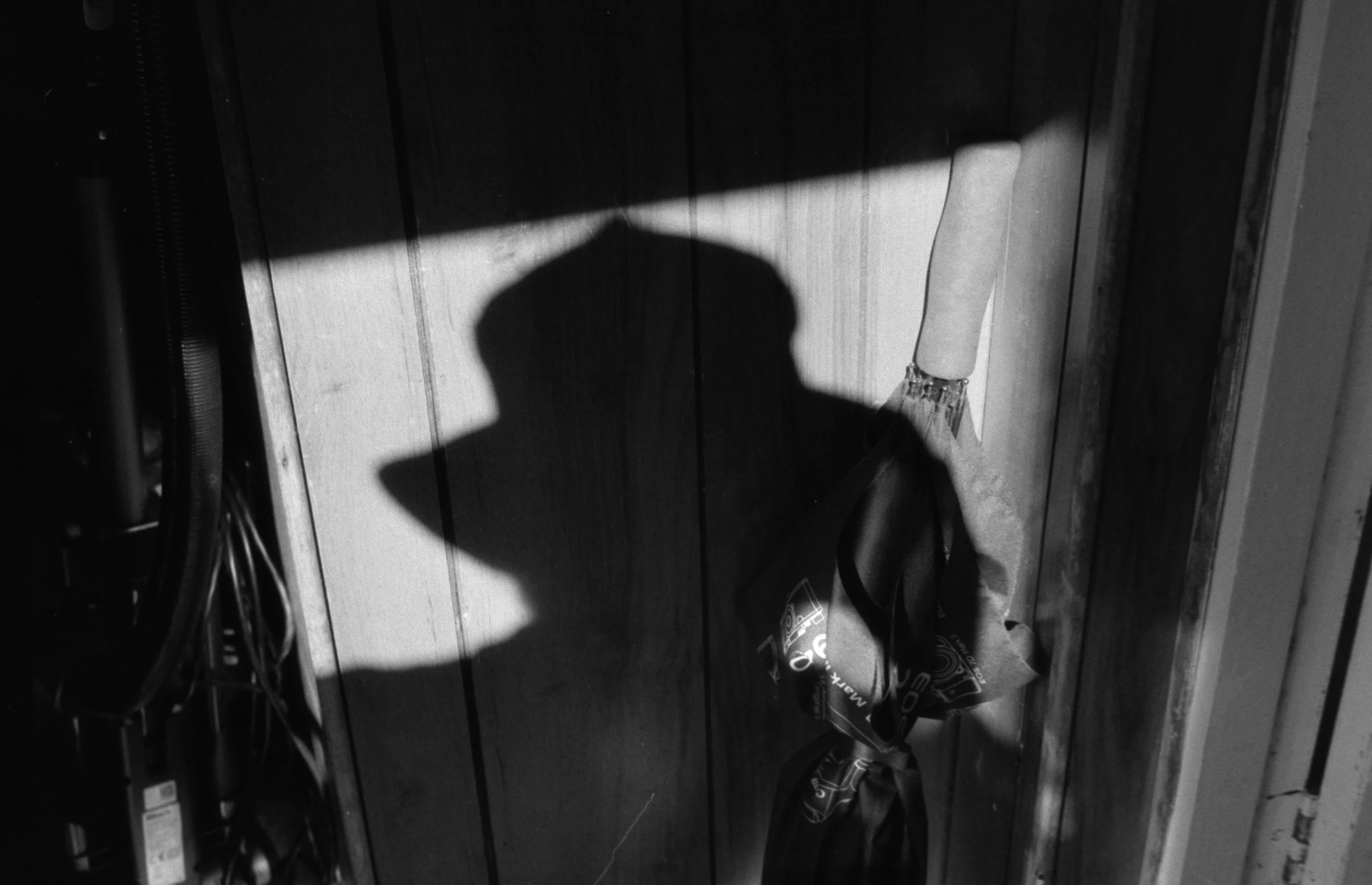 A black-and-white photo showing a silhouette of a person's profile against a wooden wall. A shadow is cast on the wall, and there are some electrical wires and a box on the left side of the image. A rolled-up item is on the right side, and a pair of 