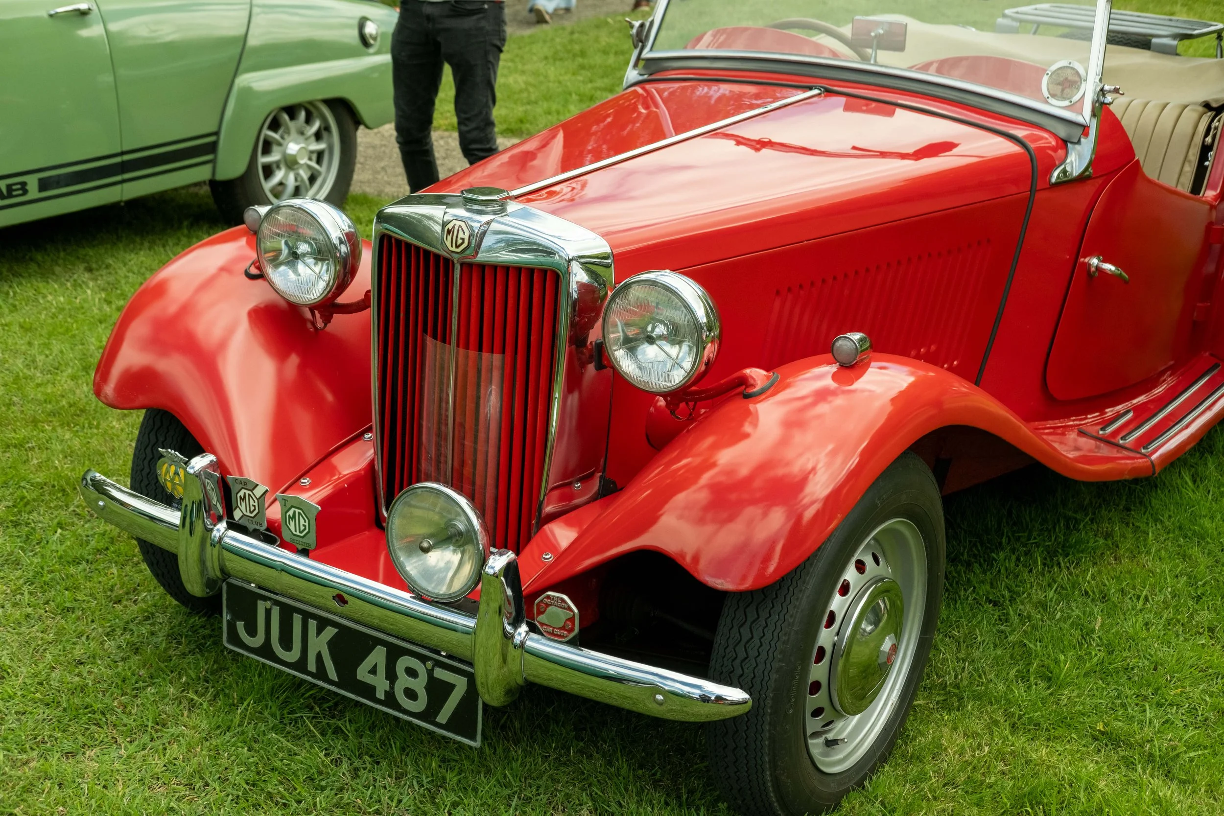 A red vintage MG car parked on grass with a black and white license plate reading 'JUK 487'. Next to it, a green car is partially visible, and a person stands behind the red car. The scene appears to be at a car show or gathering.