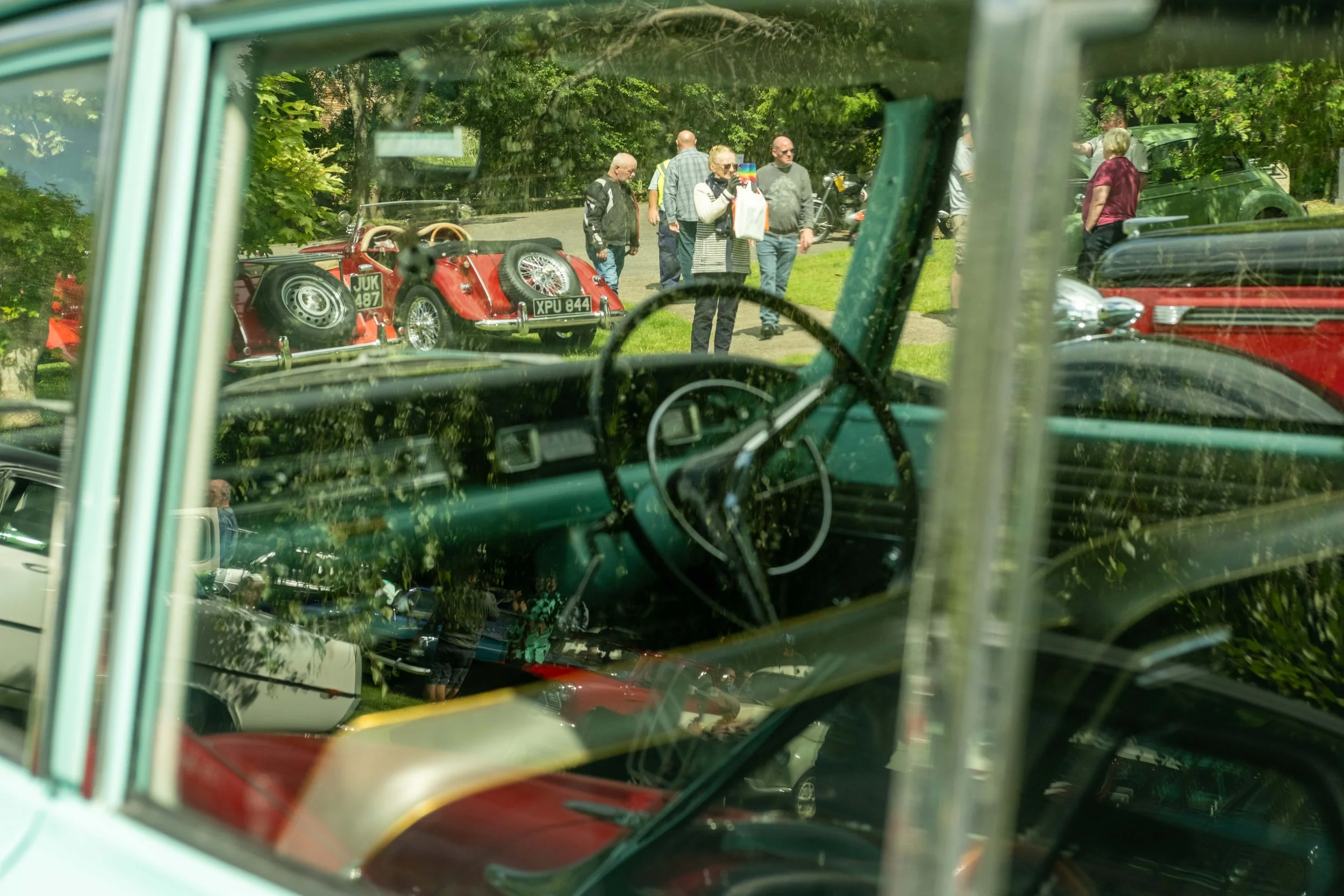 View through a vehicle window at a group of people standing in a park, with some cars including a red vintage car visible in the background. The focus is on the vintage car seen from inside another vehicle.