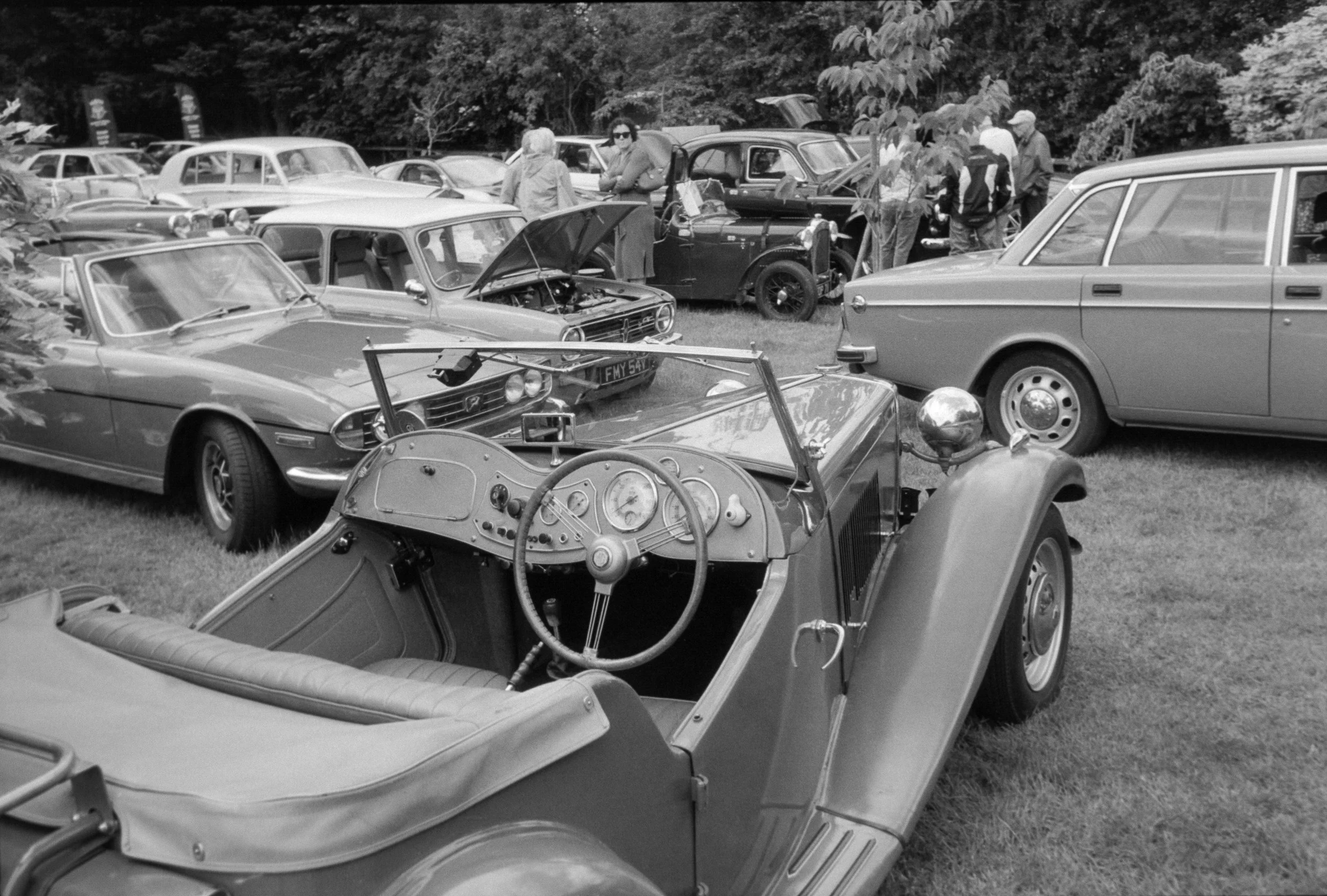 A black and white photo of a vintage car show with various classic cars parked on a grassy field. Several people are walking around, inspecting the cars, with some looking under the hoods. The background features a wooded area.