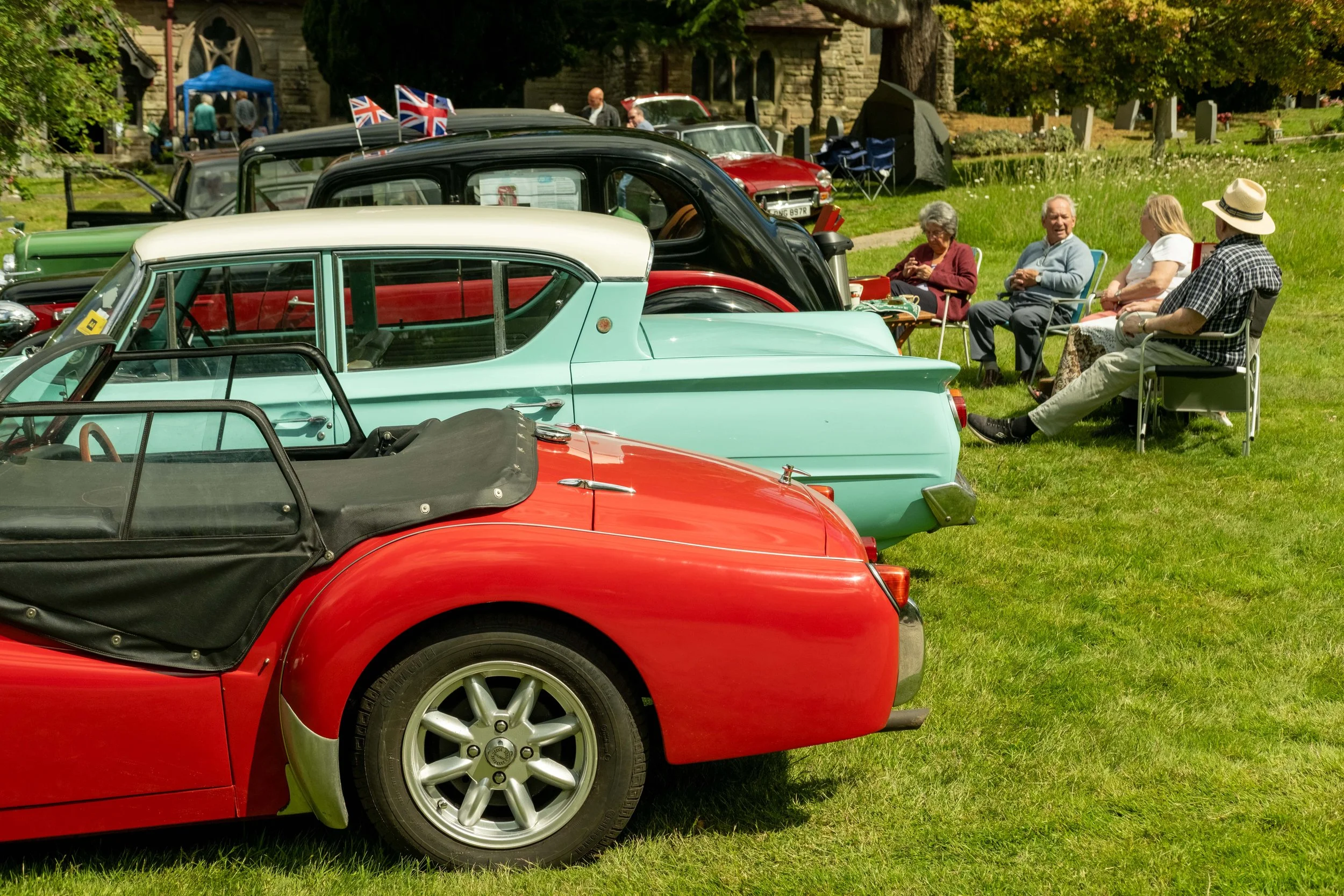 A row of vintage cars parked on grass during a car show, with four elderly people sitting and talking nearby, some with Union Jack flags on the cars.