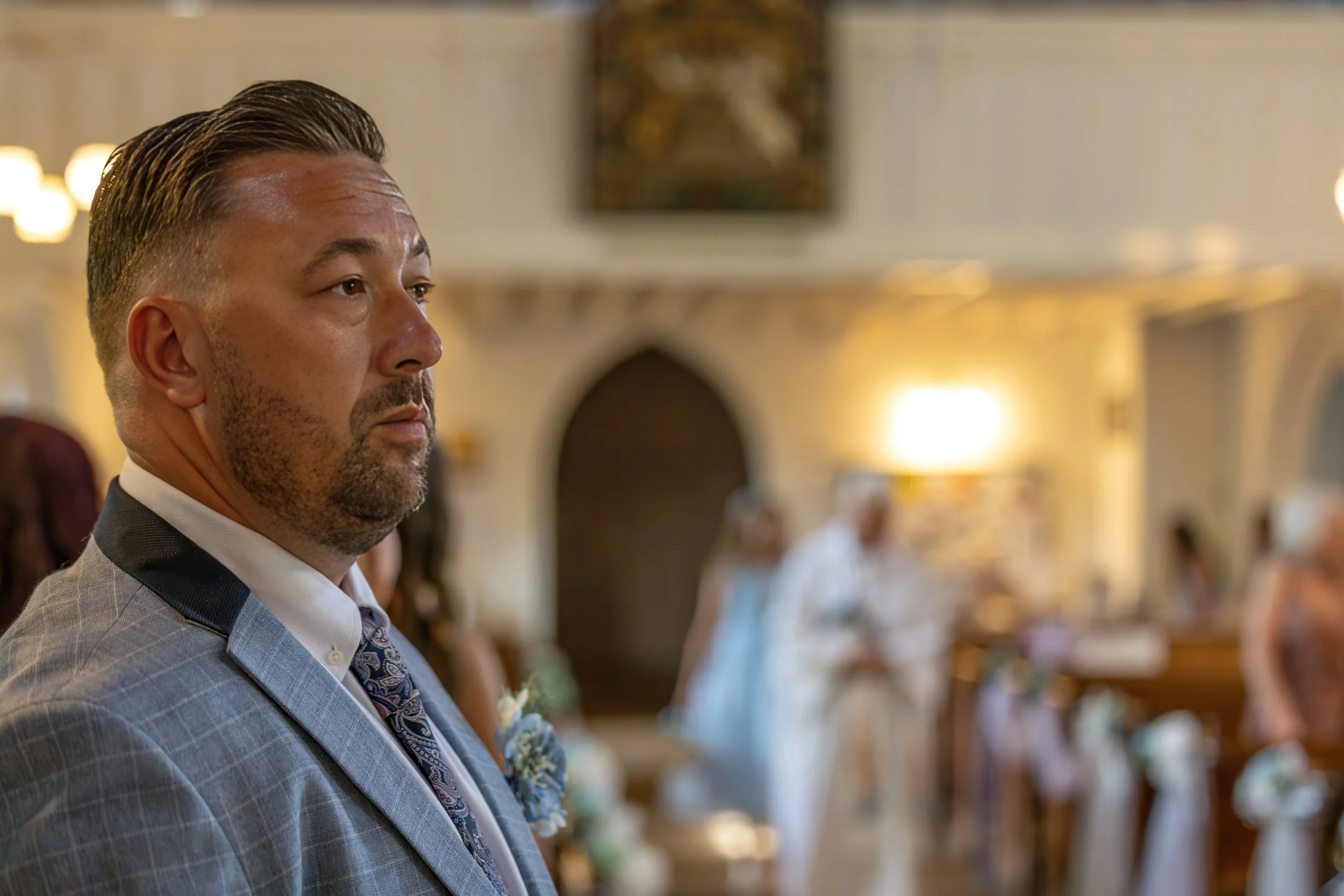 A man in a patterned suit and tie stands in a church or banquet hall during an event, with soft lighting and other guests in the background.