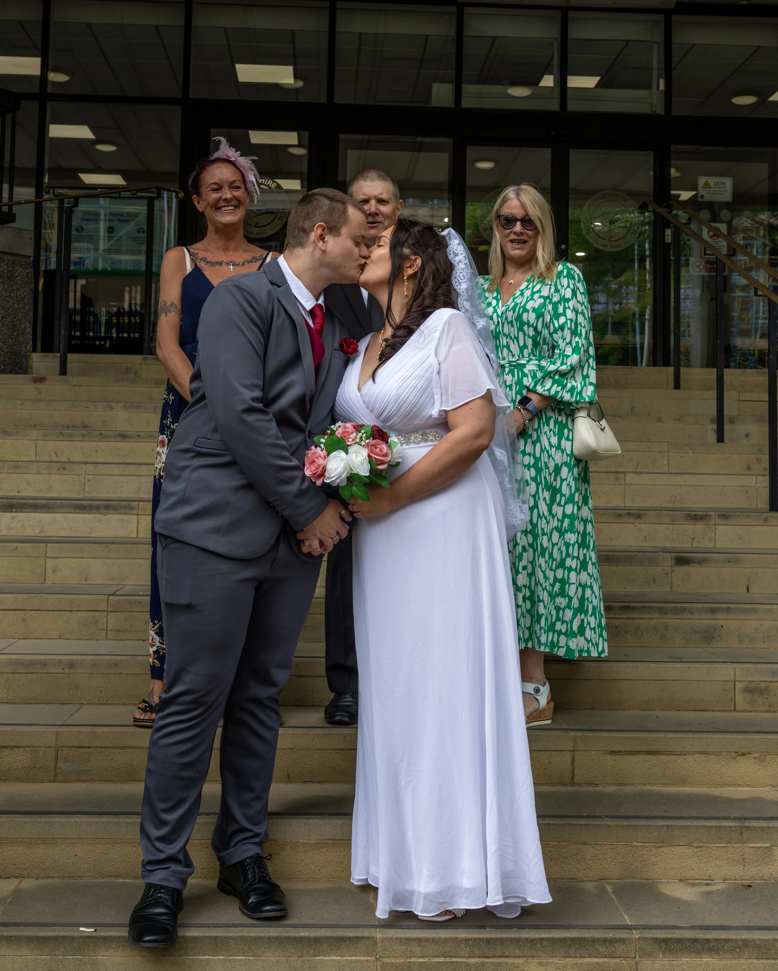 A newlywed couple sharing a kiss in front of a group of family members outside a building.
