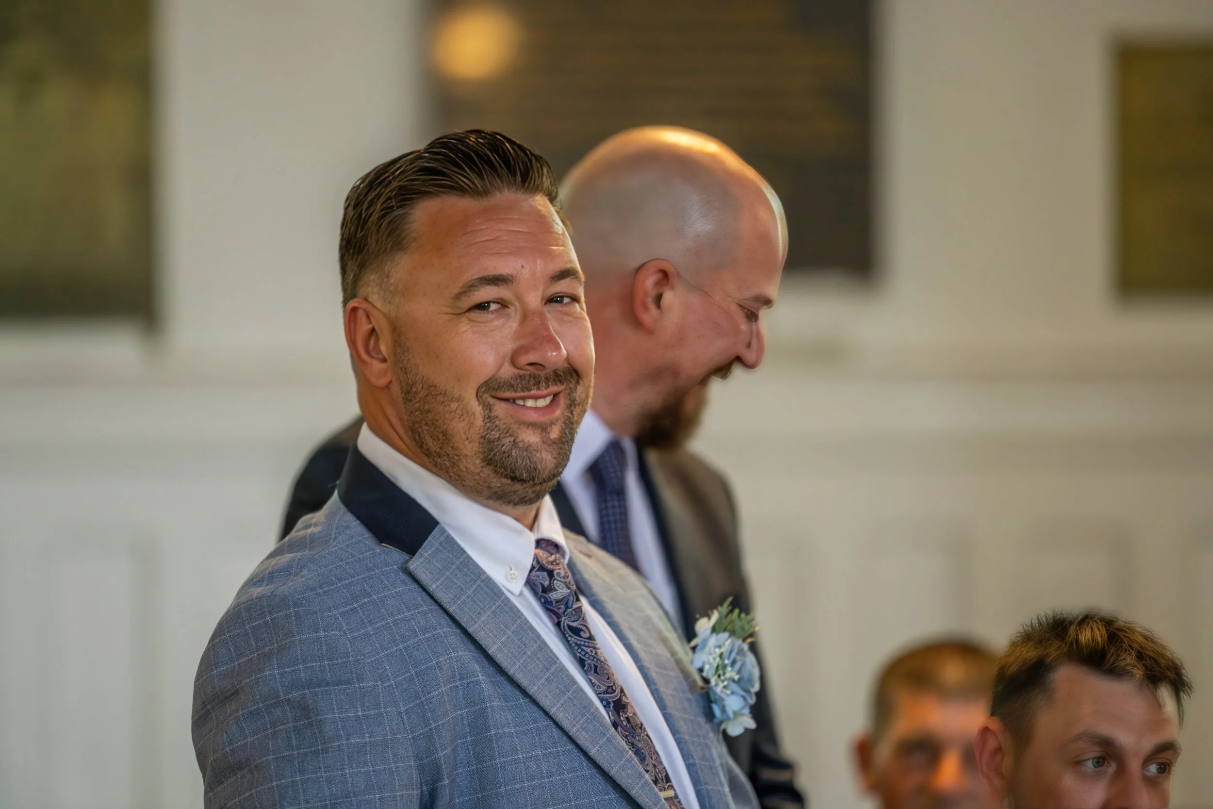 A man with dark hair and a beard, wearing a checked suit and patterned tie, smiling at a formal event, with a blurred background and another man with a shaved head and suit sitting beside him.