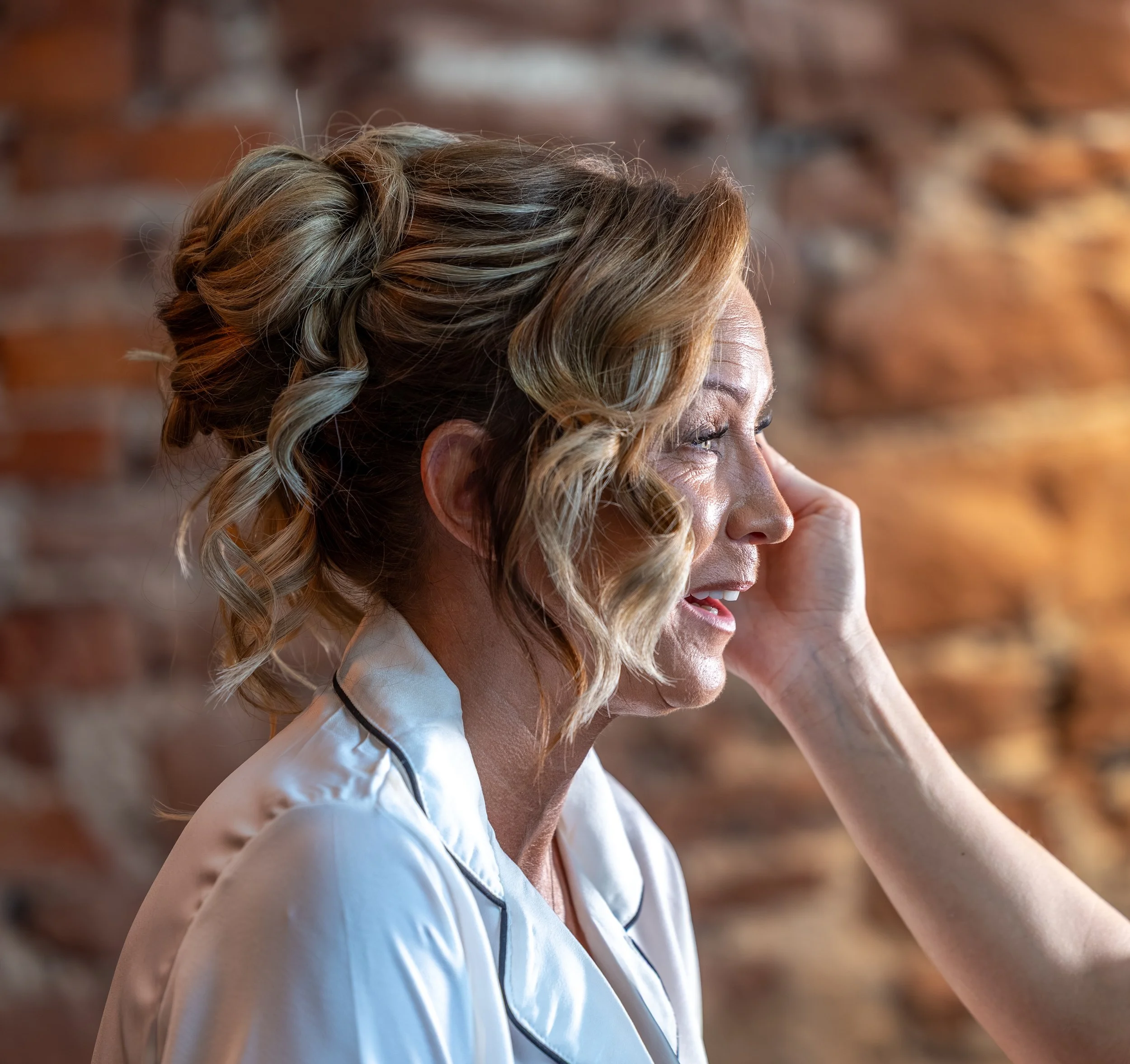 A bride with curly blonde hair styled in an updo with curls and strands falling on her shoulders, wearing a white collared shirt with black piping, seen in profile against a blurred brick wall background.