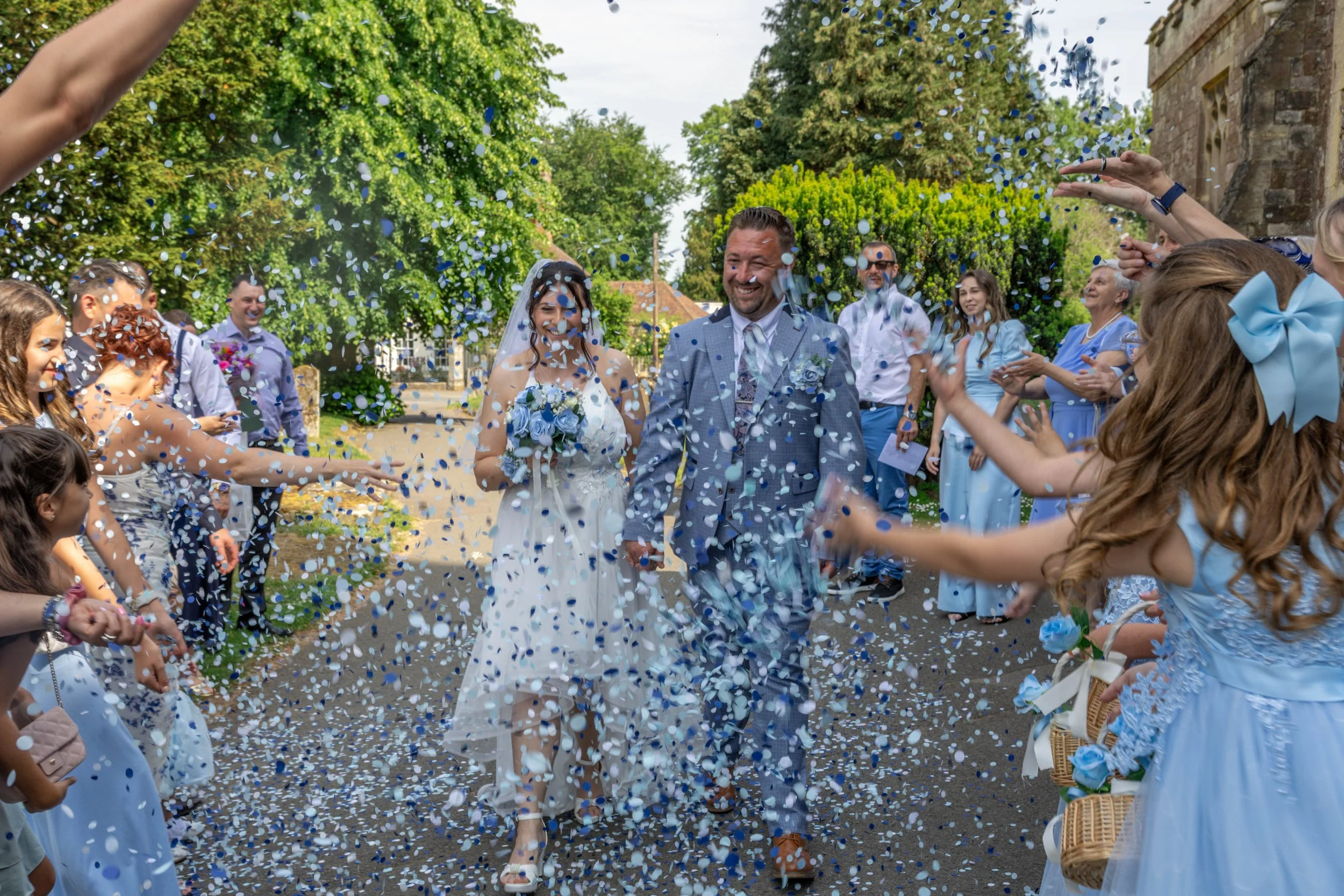 A bride and groom walking through a confetti shower during their wedding celebration while guests shower them in confetti, outdoors with green trees and a stone building in the background.