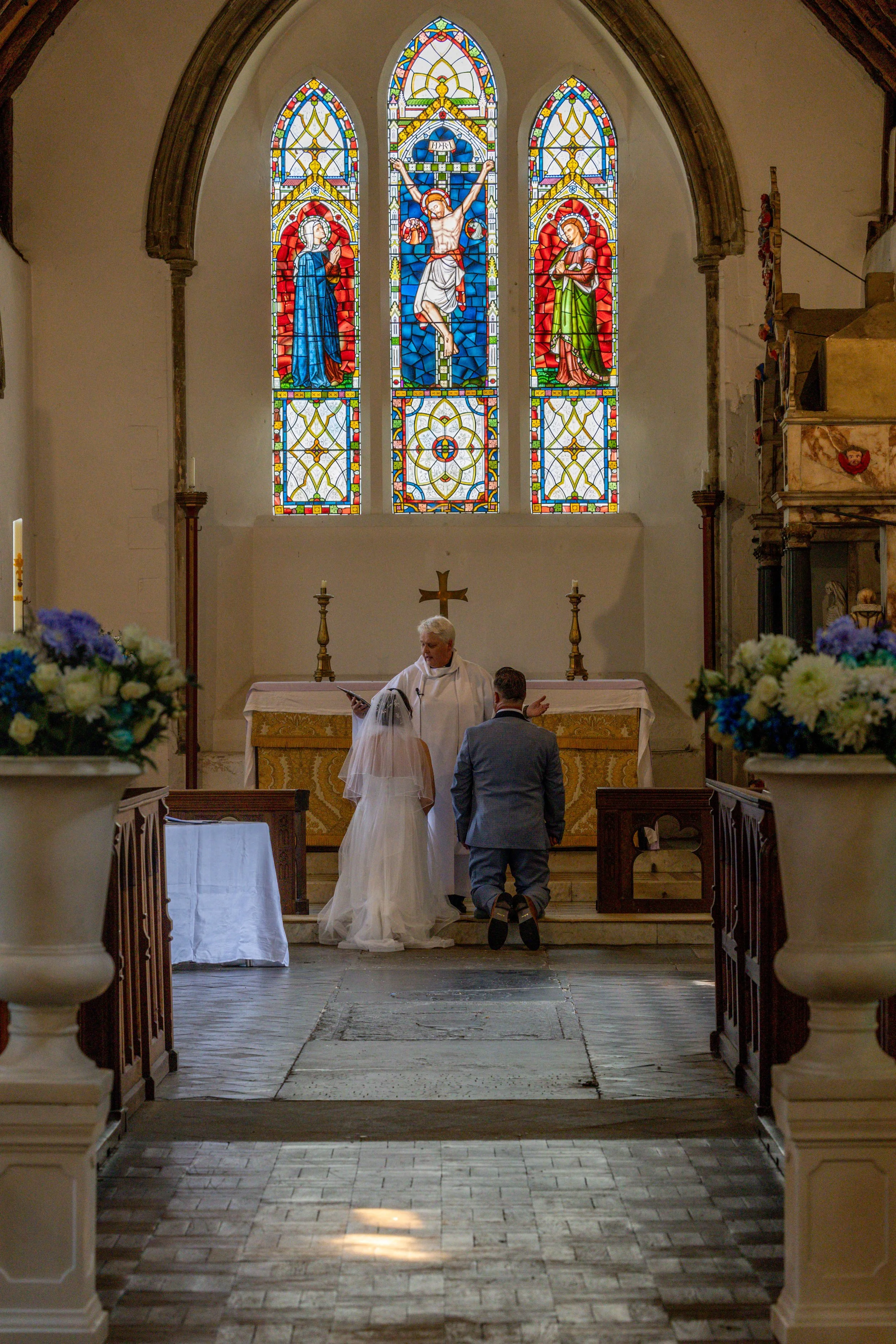 A wedding ceremony taking place in a church with stained glass windows depicting religious figures. A priest is performing the ritual in front of the bride and groom, who are kneeling.