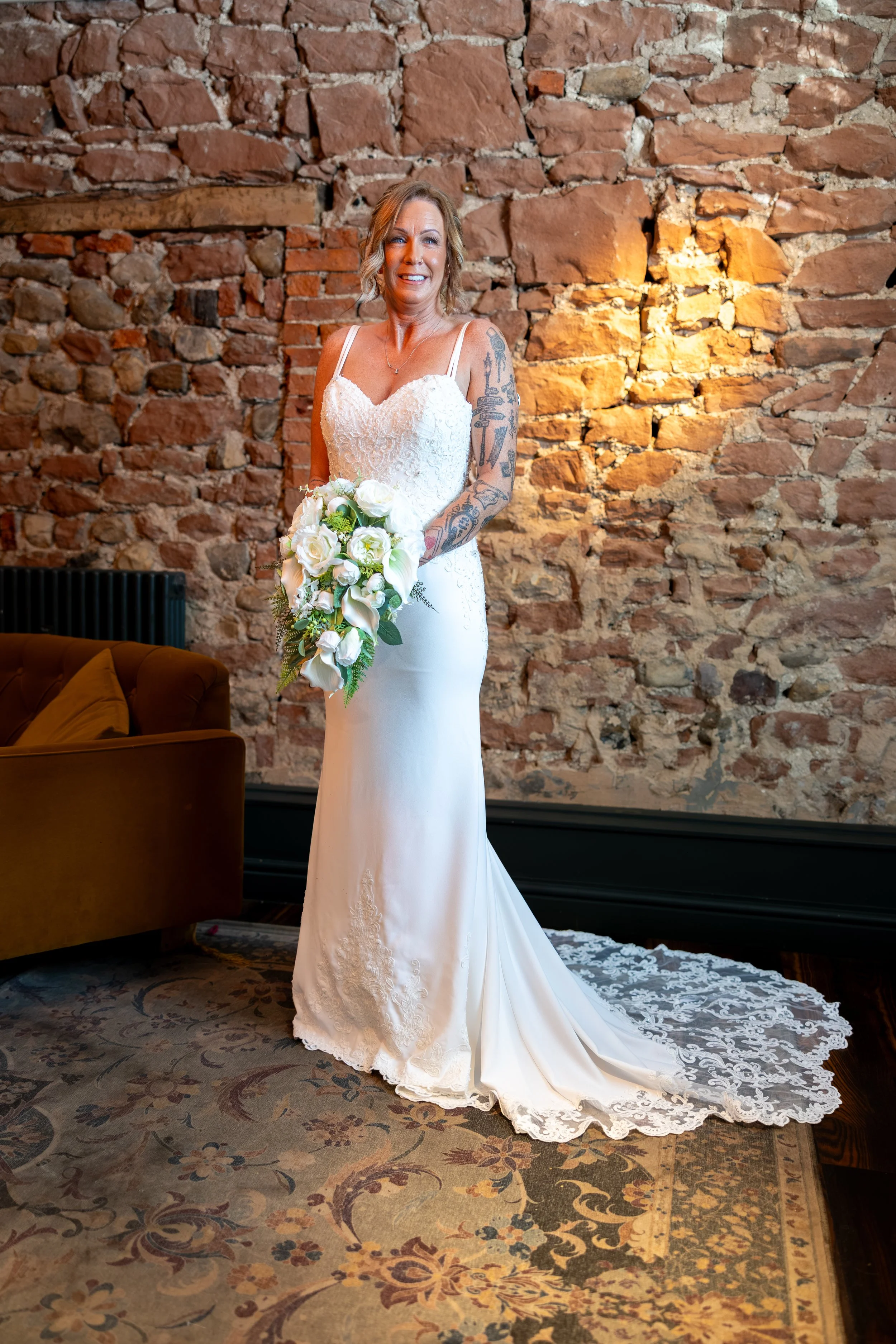 A bride in a white wedding dress holding a bouquet of white flowers, standing in front of a rustic brick wall, smiling.