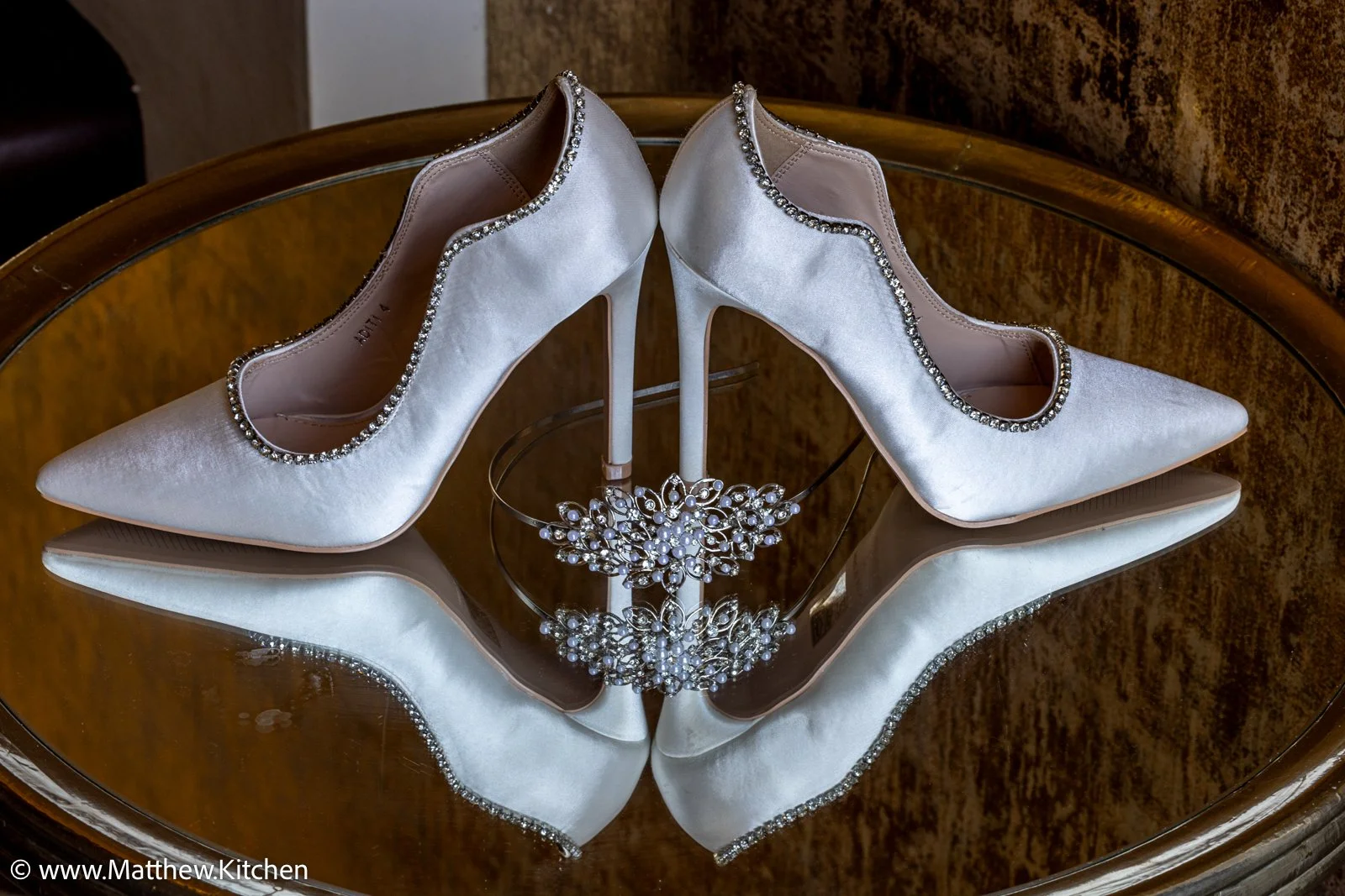 A pair of white satin high-heeled shoes with rhinestone trim on a mirrored surface, with a matching tiara and necklace reflected underneath.