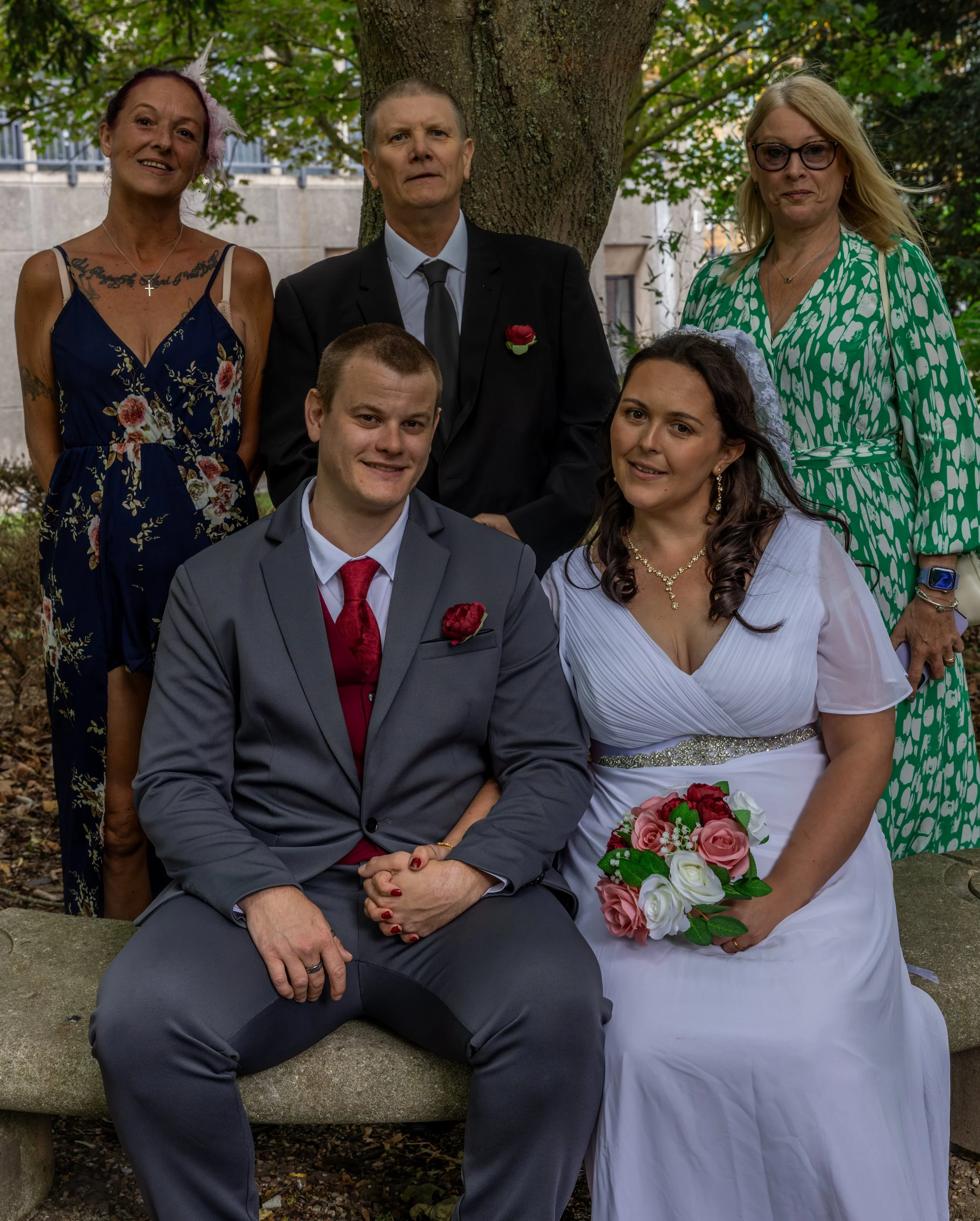 Group of six people outdoors, one woman in a white wedding dress holding a bouquet, others in formal attire, standing and sitting near a tree, background includes a stone wall and trees.