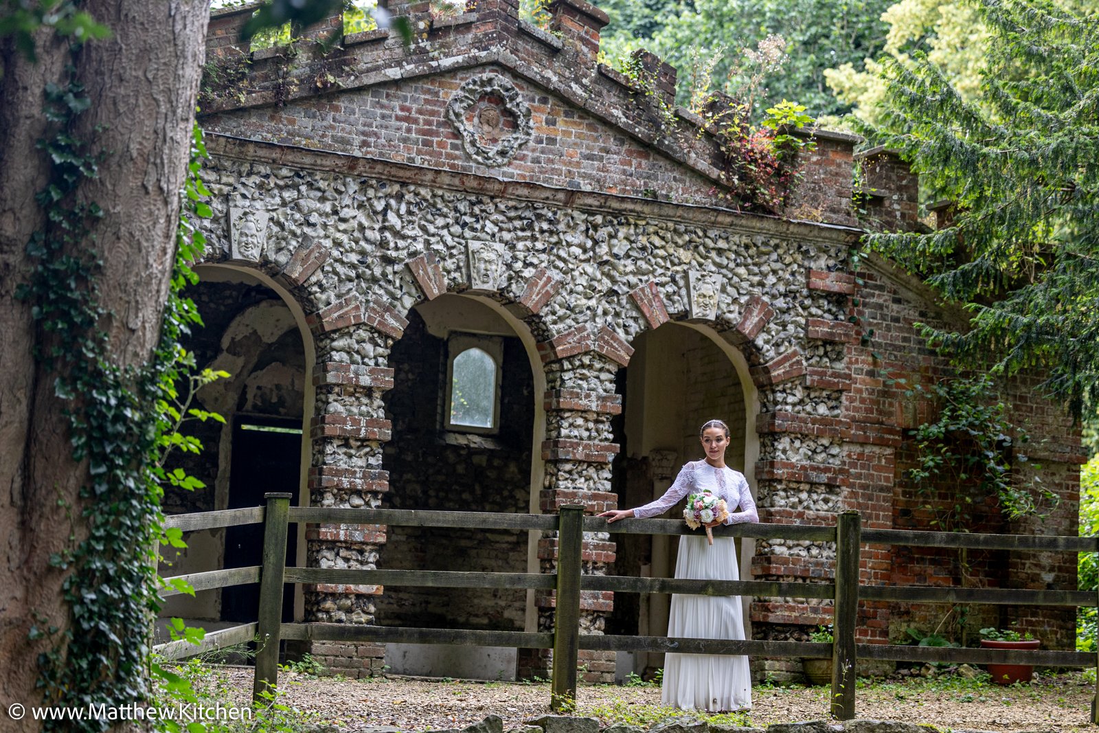 A woman in a white dress holding a bouquet of flowers standing outdoors near an old brick and stone building with arched openings, surrounded by trees.