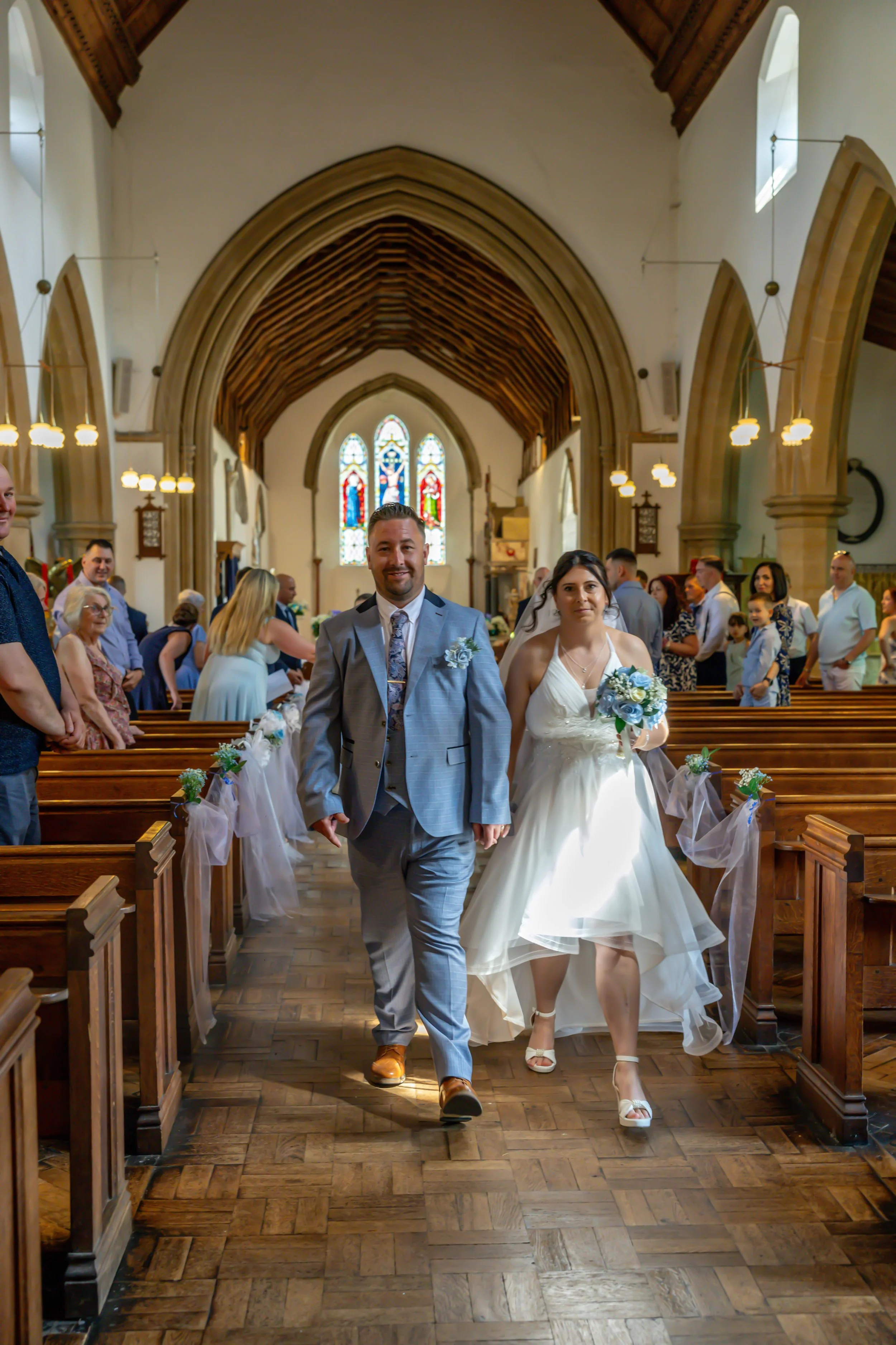 A bride and groom walking down the aisle inside a church during their wedding ceremony, with guests seated on either side and a stained glass window at the front.