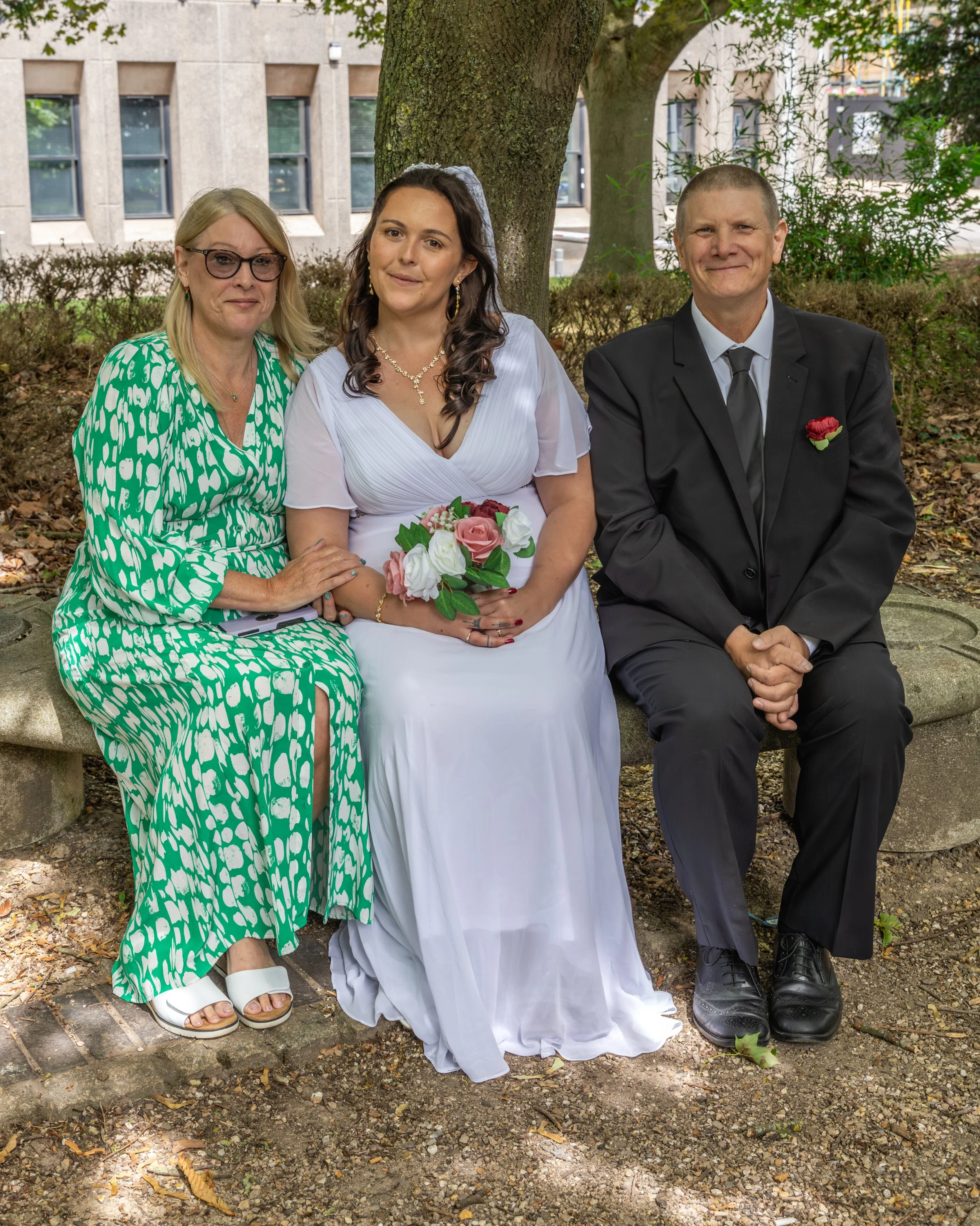 Three people sit on a bench outdoors beneath a tree. The middle person is a woman in a white wedding dress holding a bouquet of pink and white roses. To her left is an older woman with blonde hair, glasses, and a green patterned dress. To her right i