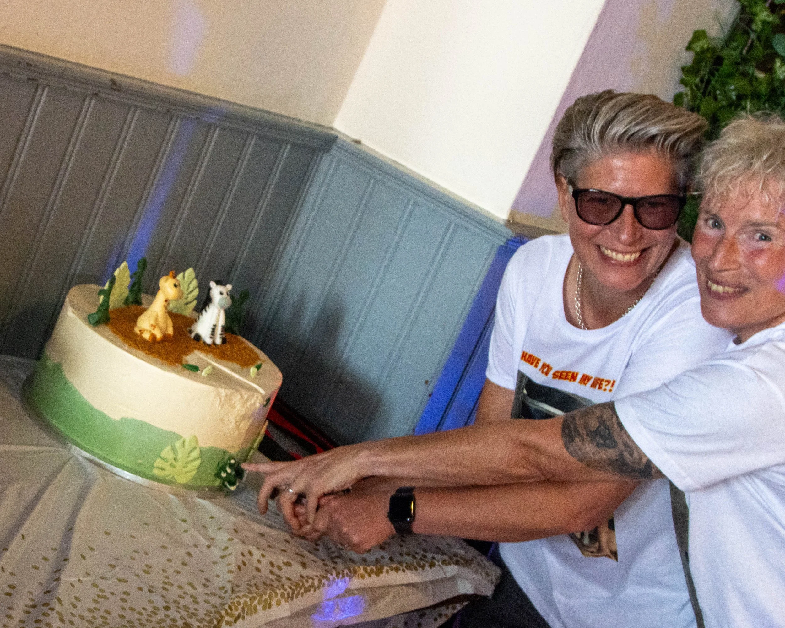 Two women smiling and cutting a large birthday cake decorated with jungle animals, with a giraffe and zebra on top, and green leaves around the sides.