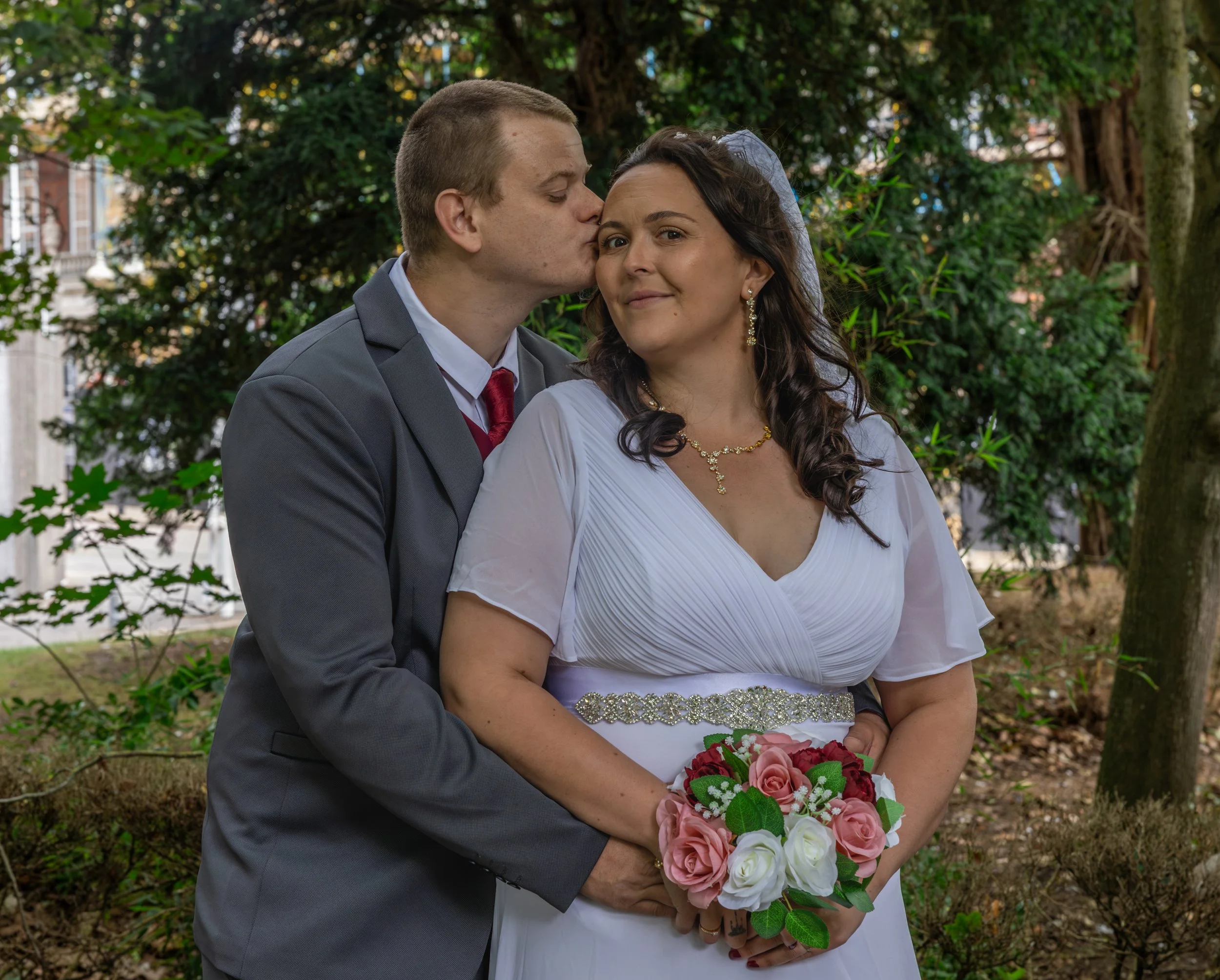 A newlywed couple standing outdoors. The man is kissing the woman on her forehead. The woman is holding a bouquet of pink and white roses. They are dressed in wedding attire with the man in a gray suit and red tie, and the woman in a white wedding dr