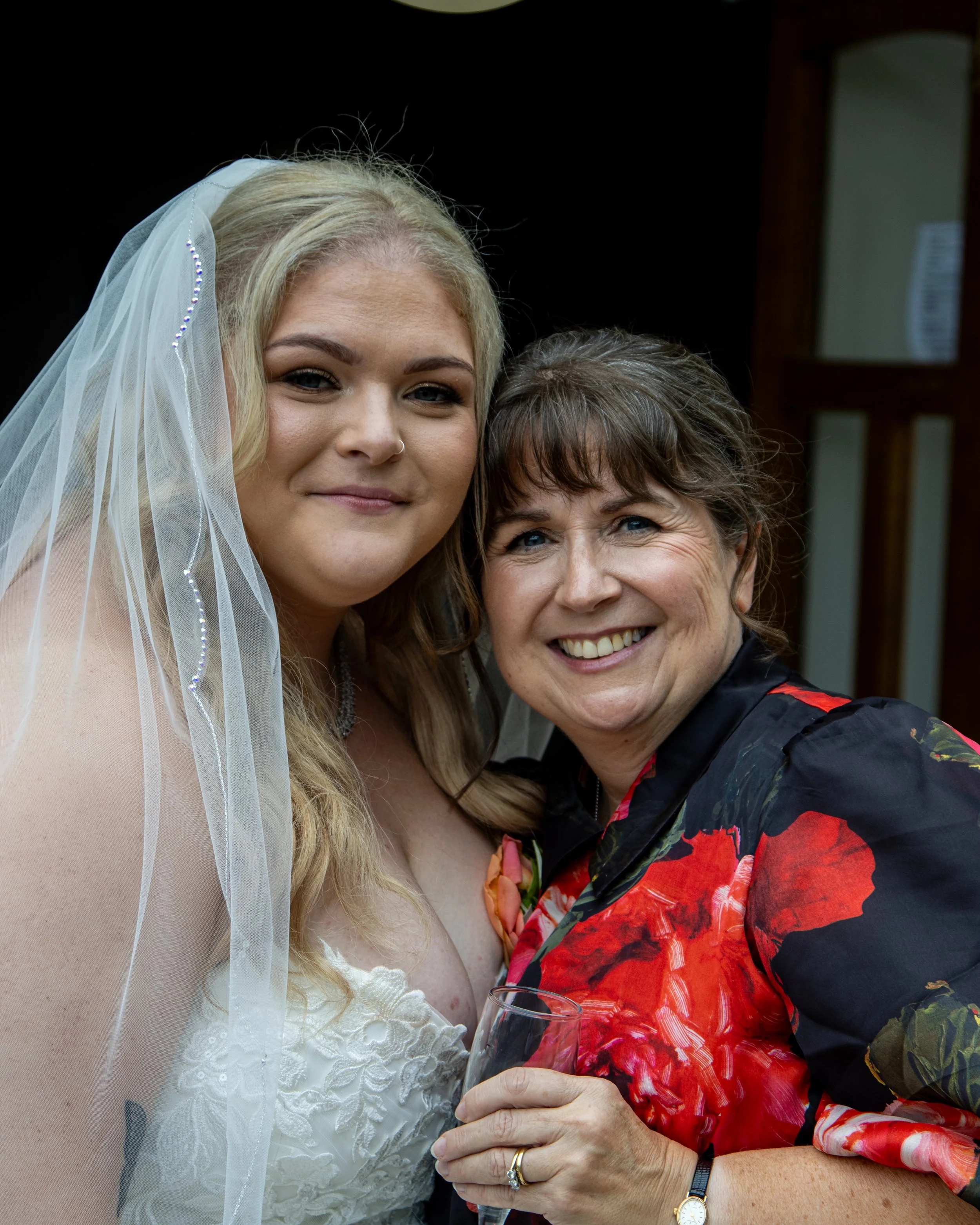 Two women smiling, one wearing a wedding dress with a veil, and the other in a floral blouse holding a glass of wine.
