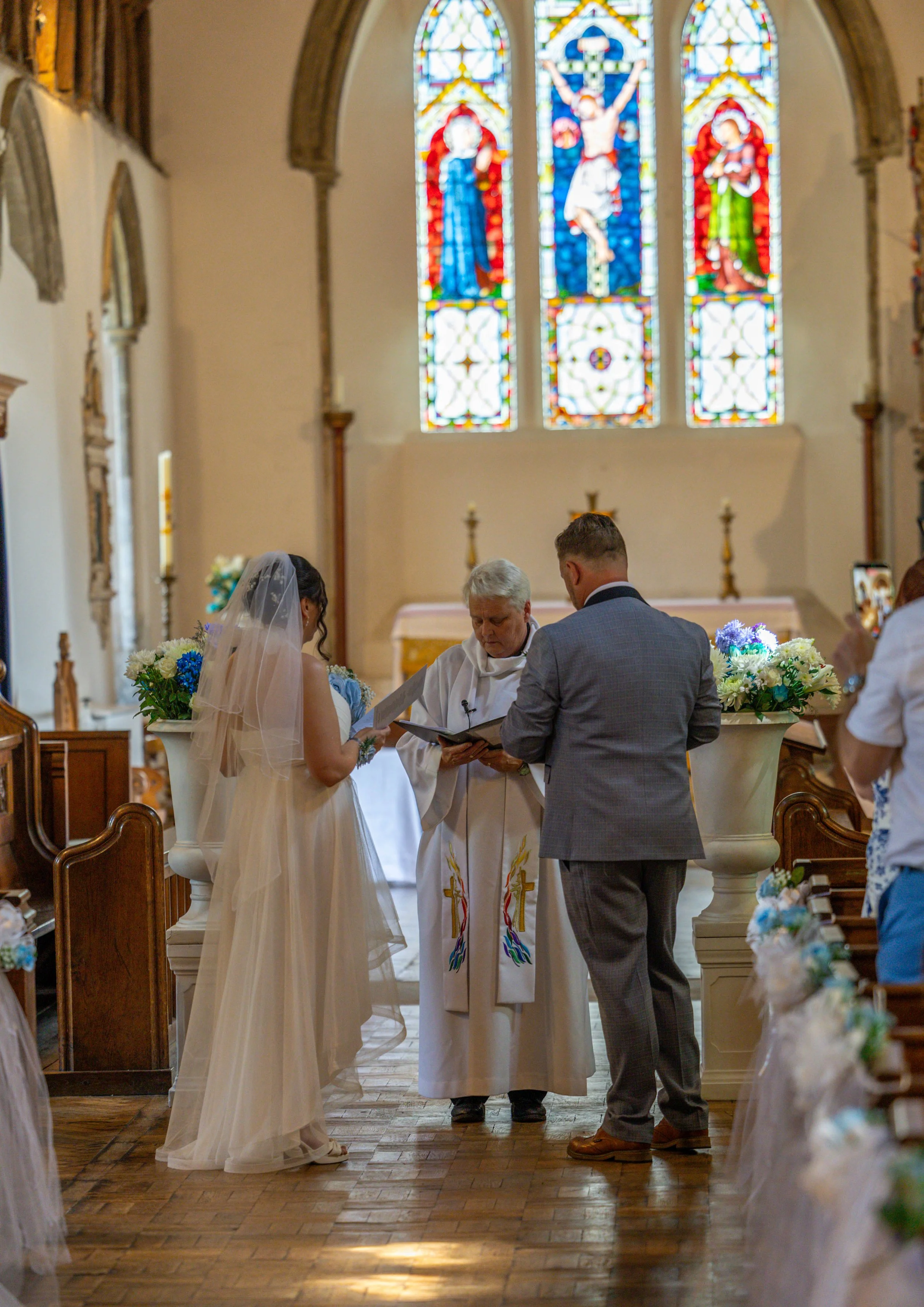 A bride and groom standing before a priest during a wedding ceremony in a church with stained glass windows.