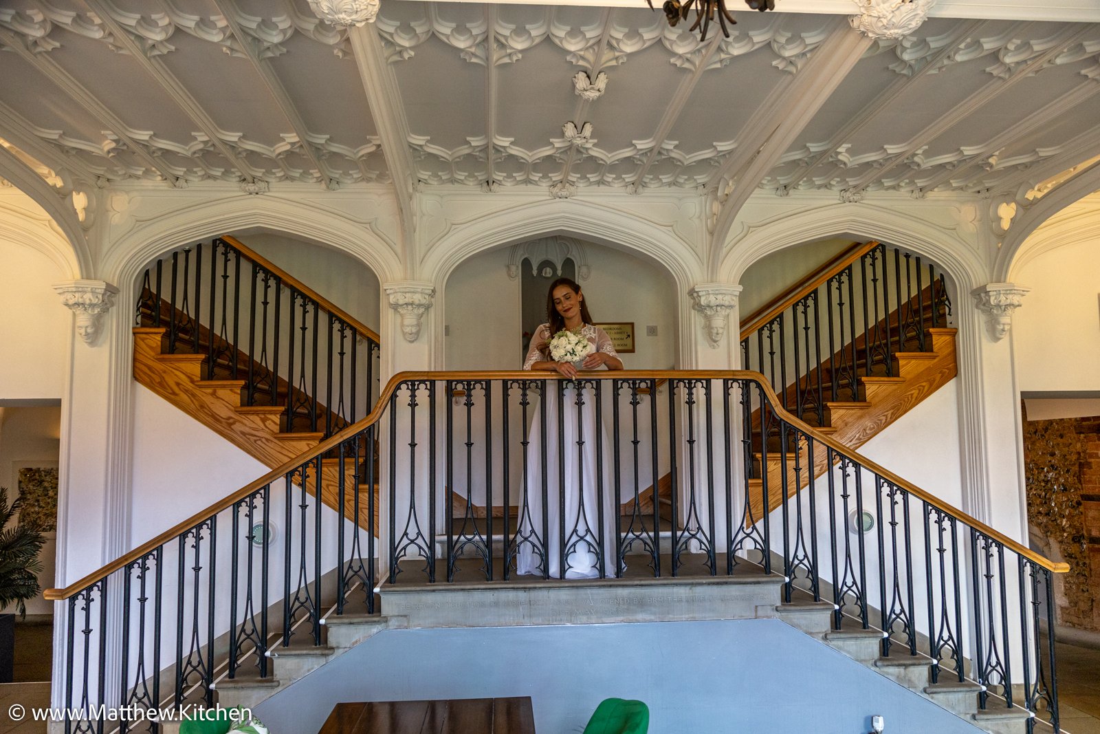 A bride holding a bouquet stands on a balcony inside a building with ornate white ceiling details and wooden stairs on either side.
