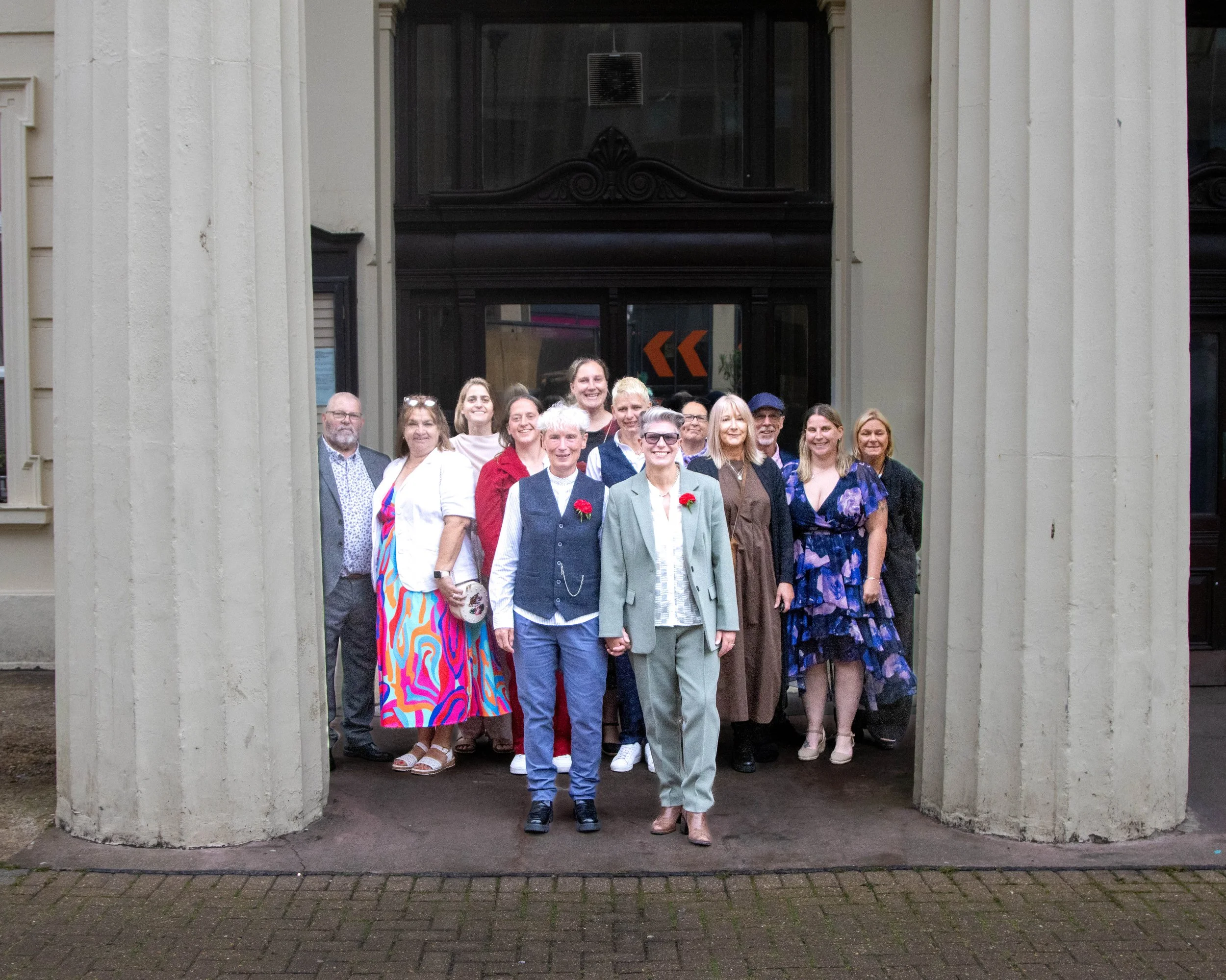 Group of people standing together outdoors in front of a building, smiling for the camera. Some individuals are wearing formal attire, and a few have red flowers pinned to their clothing.