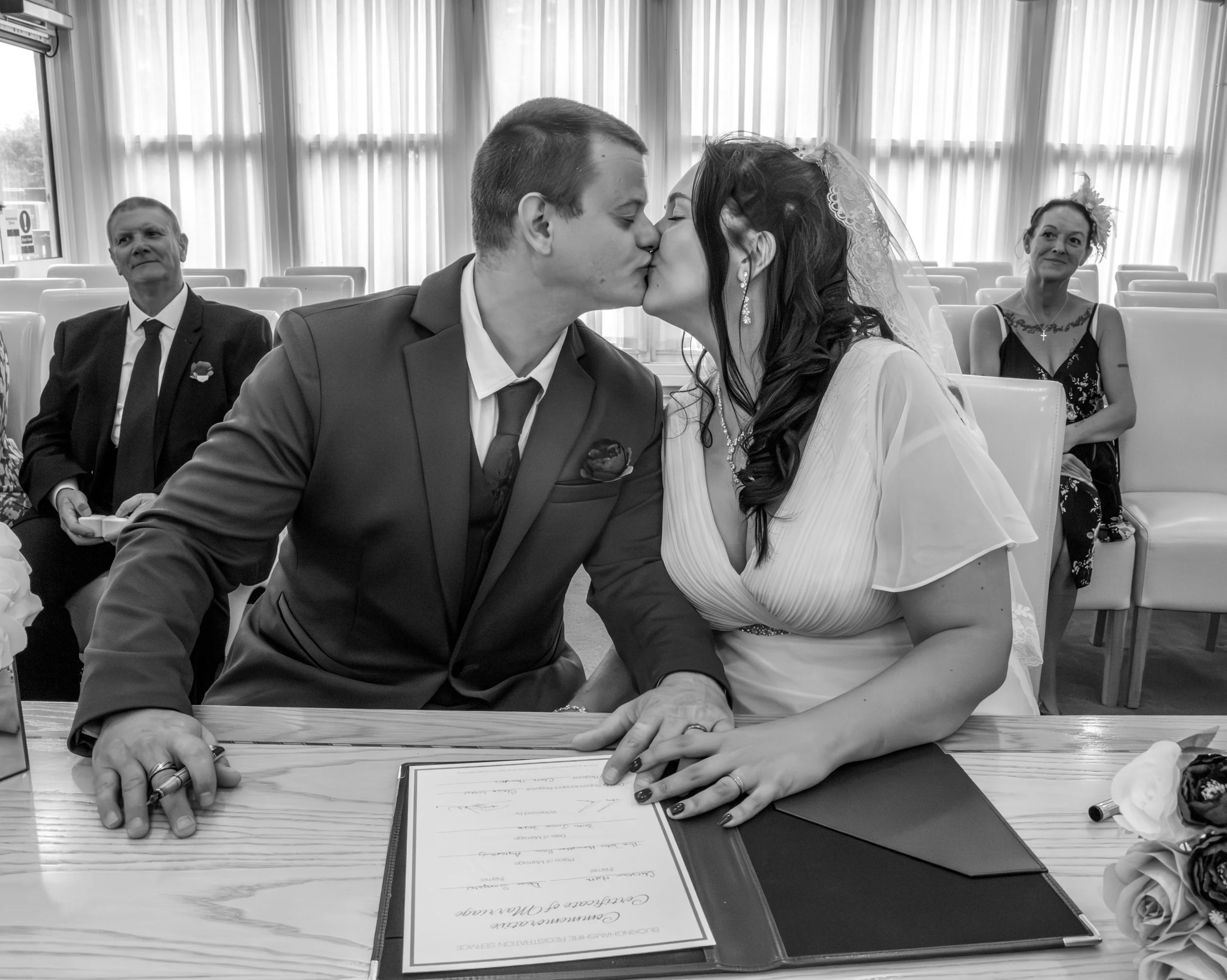Black and white photo of a wedding ceremony with a couple kissing, two people sitting behind them, a man in a suit and a woman in a dress with a floral headpiece, wedding document on the table.
