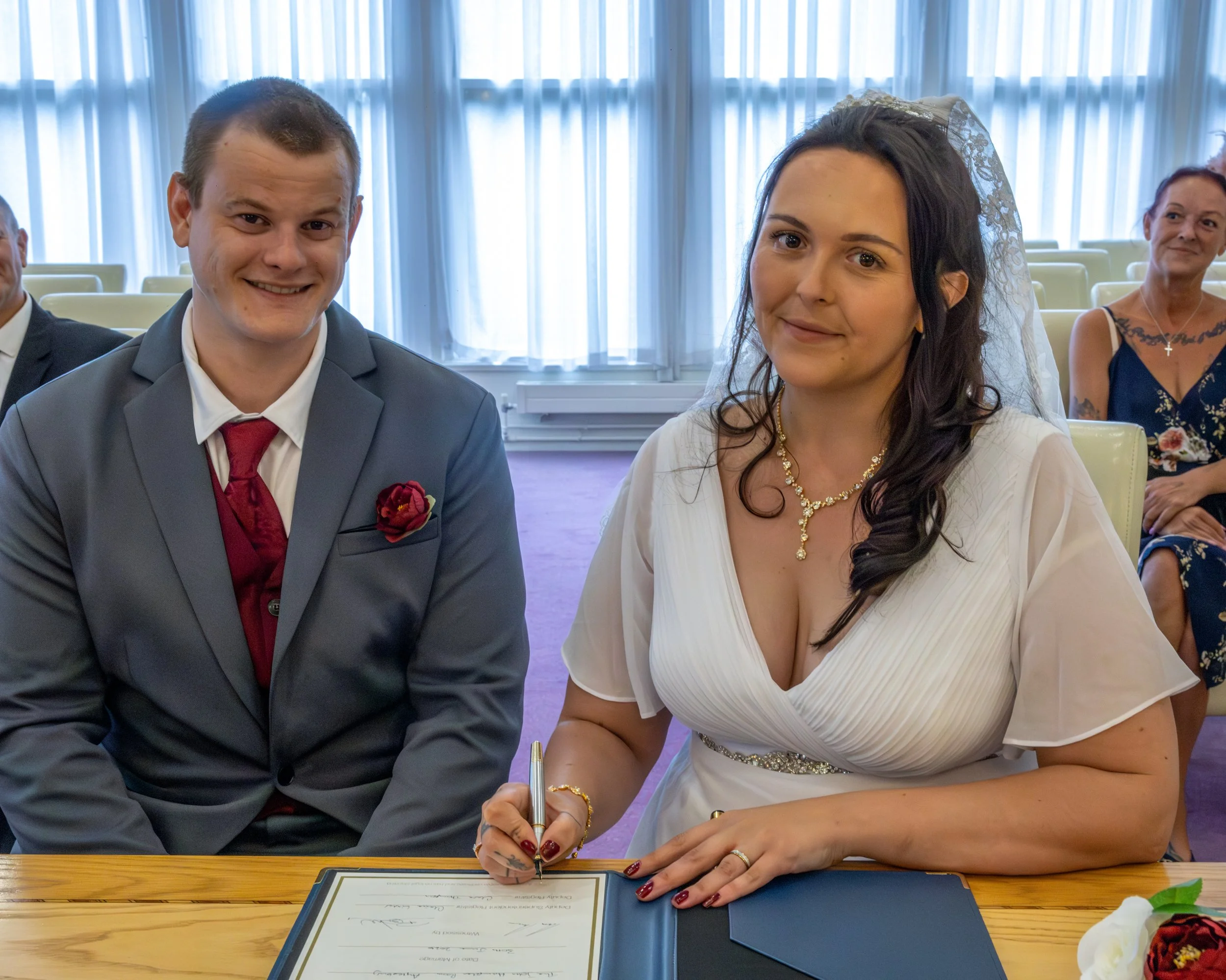 A bride and groom at a wedding ceremony seated at a table, with the bride signing a wedding document. The groom is smiling and dressed in a gray suit with a red tie and boutonniere. The bride has long dark hair, wears a white dress, jewelry, and a la