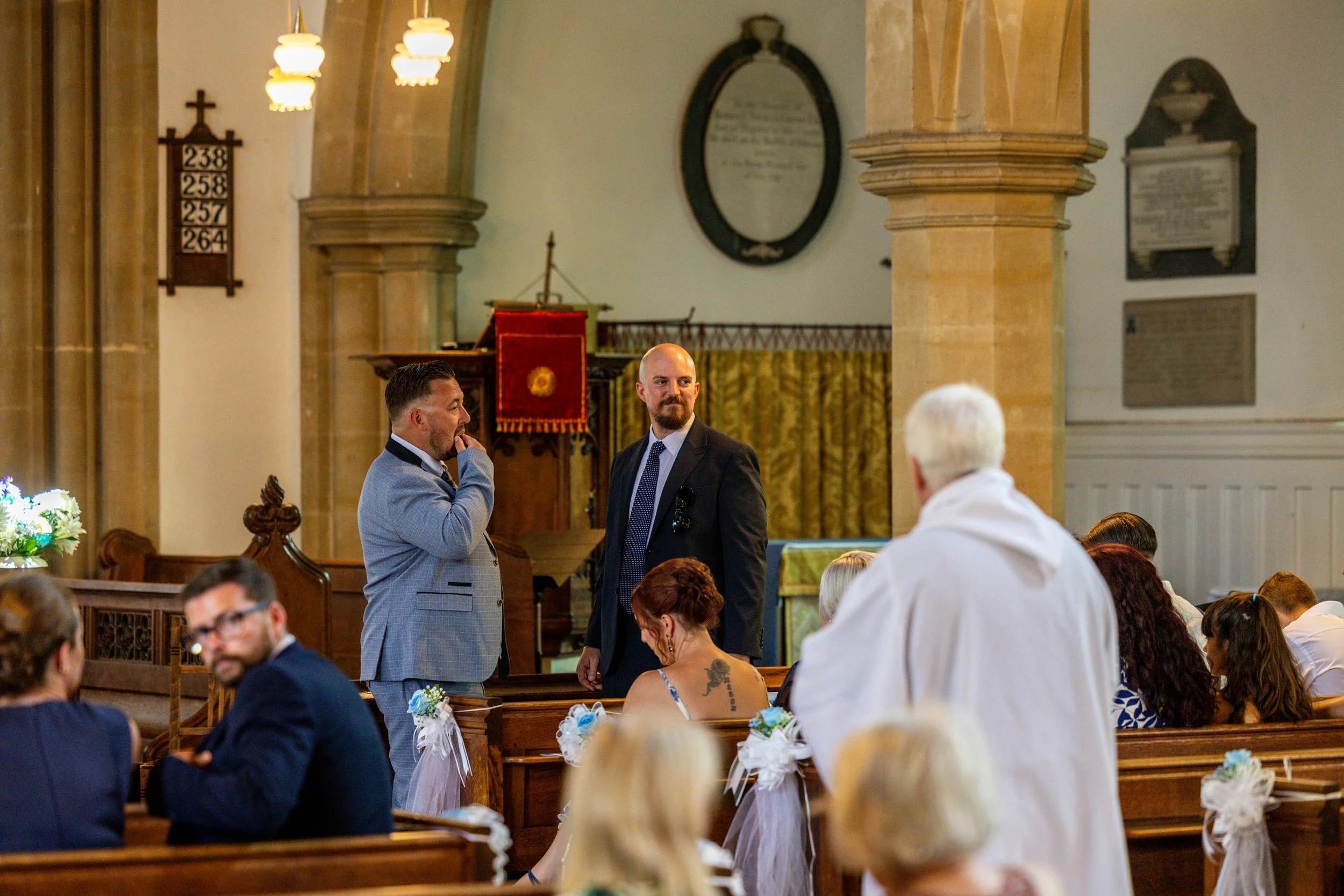 Two men dressed in suits are standing inside a church, engaging in conversation. The church interior has wooden pews decorated with white and blue ribbons, and plaque and memorials on the walls.