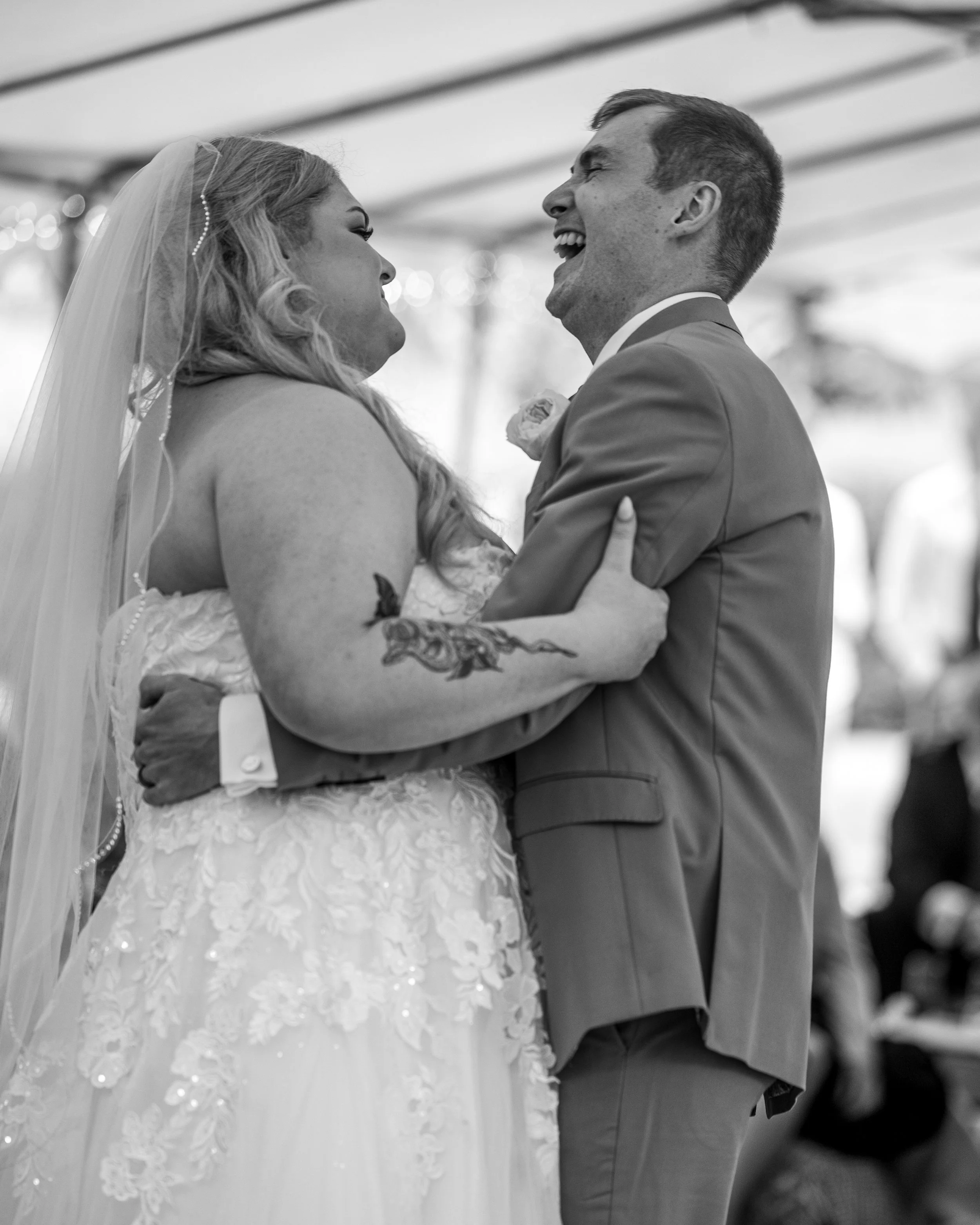 A black and white photo of a bride and groom smiling and looking at each other, sharing a dance at their wedding.