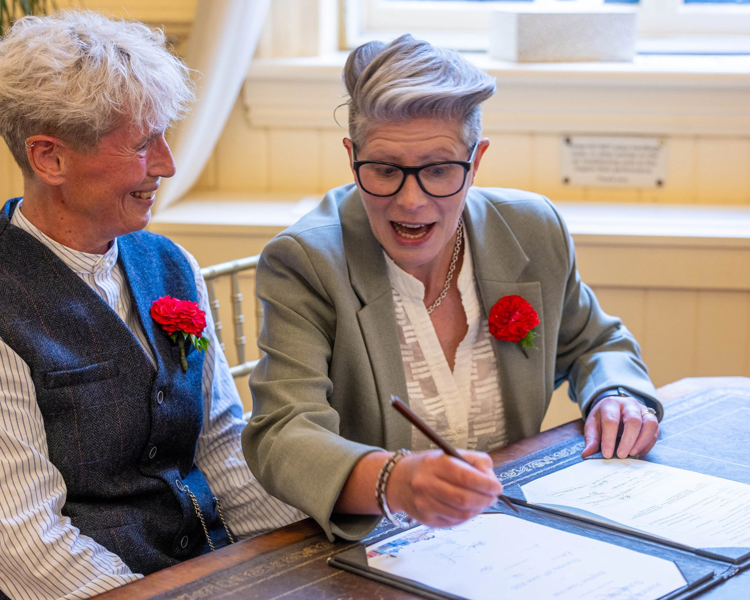 Two women sitting at a table, one signing a document and both wearing red flower boutonnieres, in a warmly lit room with wood paneling and a window in the background.