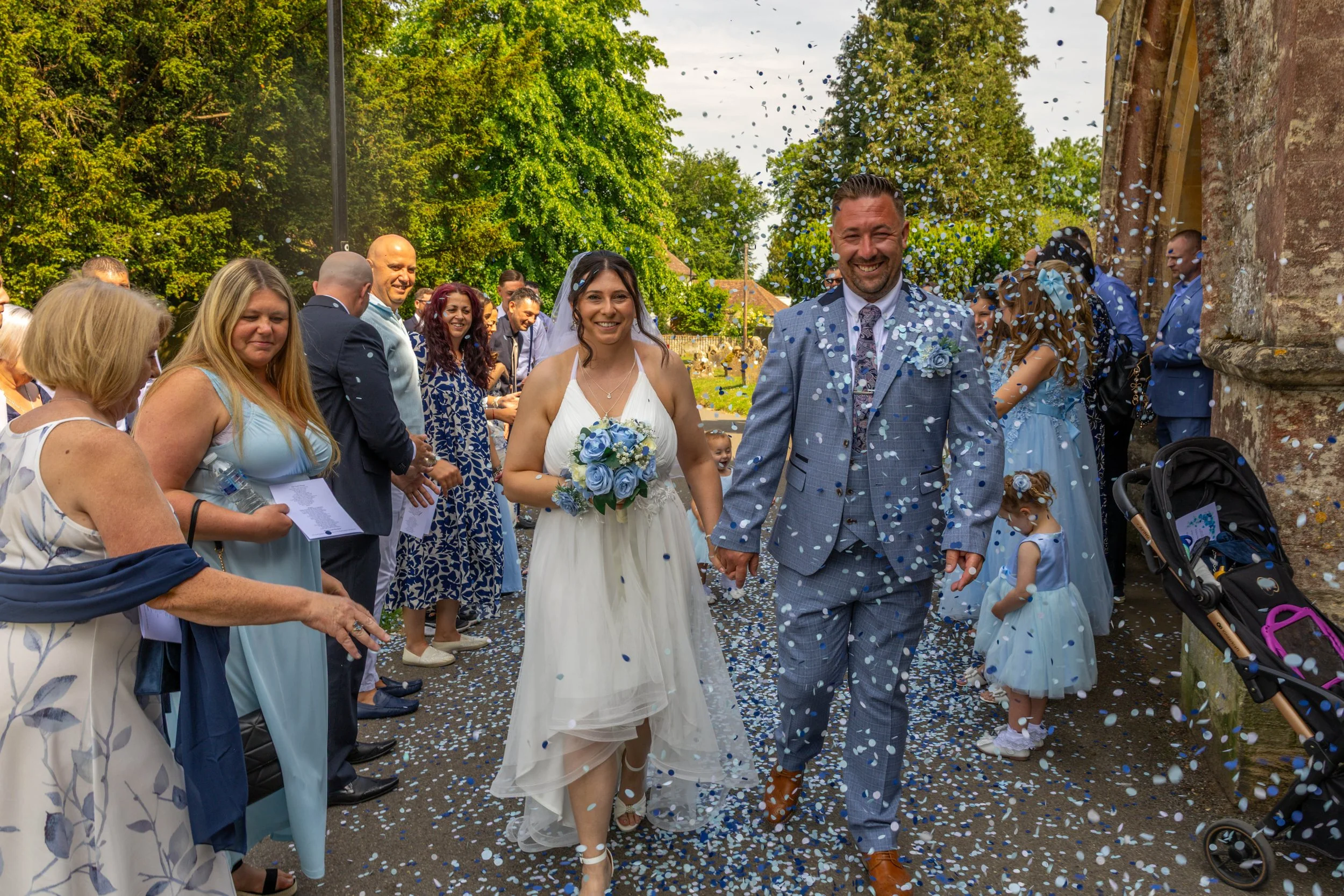 A newlywed couple holding hands and smiling as they walk through a confetti shower outdoors. The bride wears a white dress with a bouquet, and the groom wears a gray plaid suit. Guests surround them, celebrating near a church with trees in the backgr