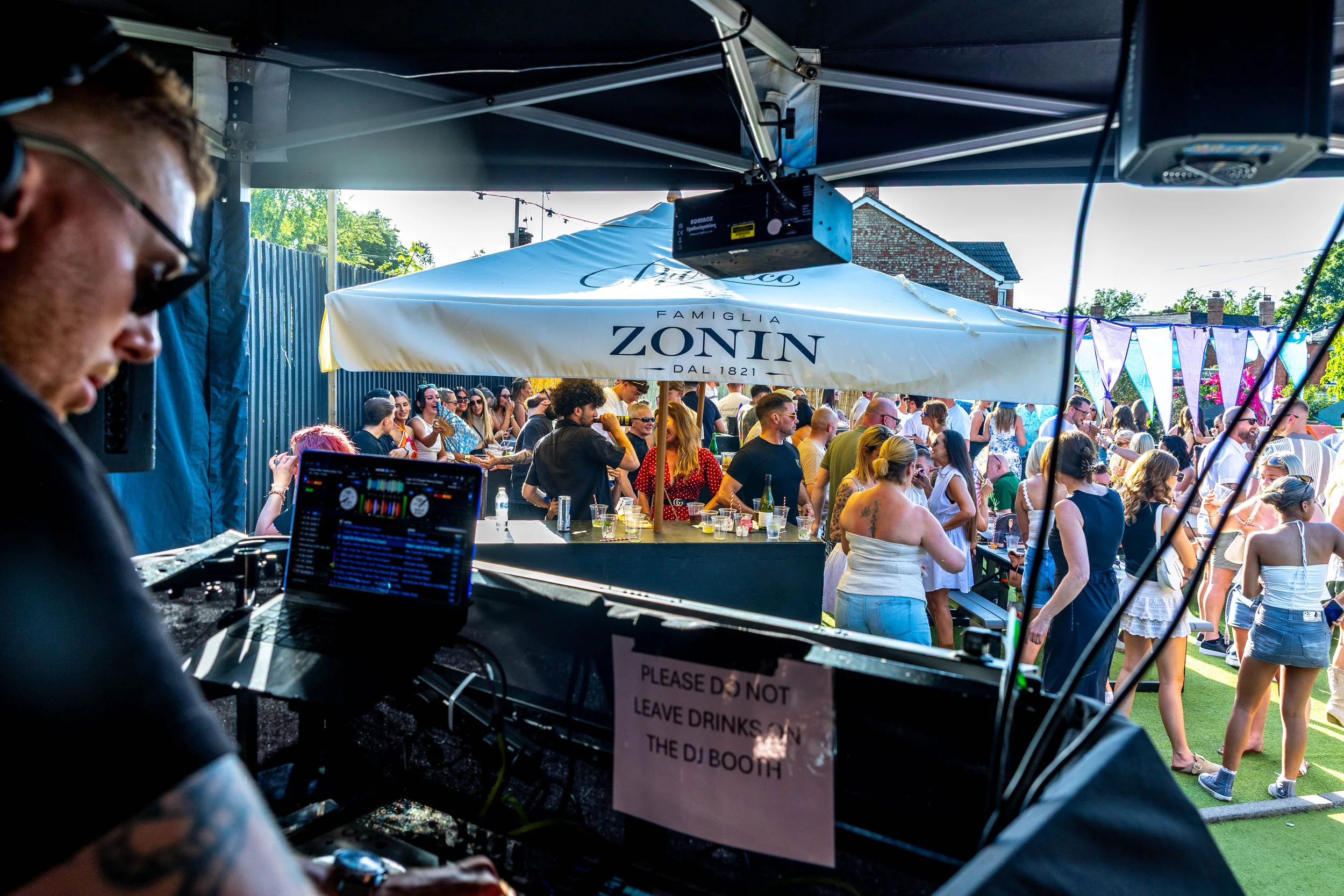 A DJ is working at a booth outdoors during a lively party with many people dancing and socializing under umbrellas, with a house and trees in the background.