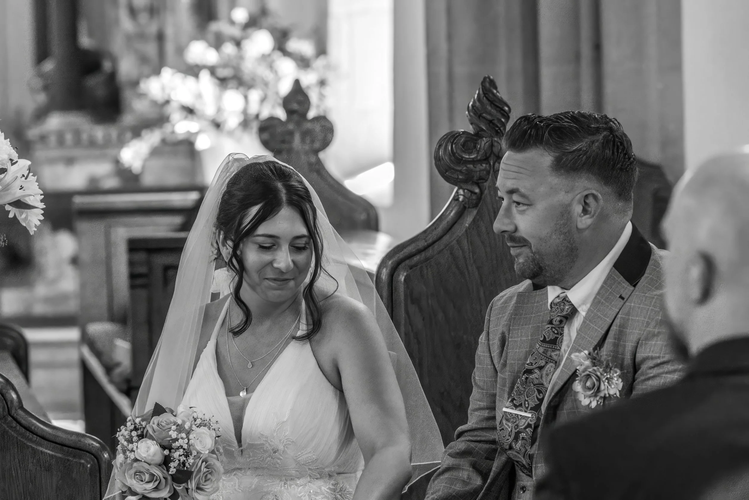 A bride and groom sitting together inside a church during their wedding ceremony, the bride is holding a bouquet of flowers and looking down, while the groom looks at her. They are seated on ornate wooden chairs with a religious statue and a decorate