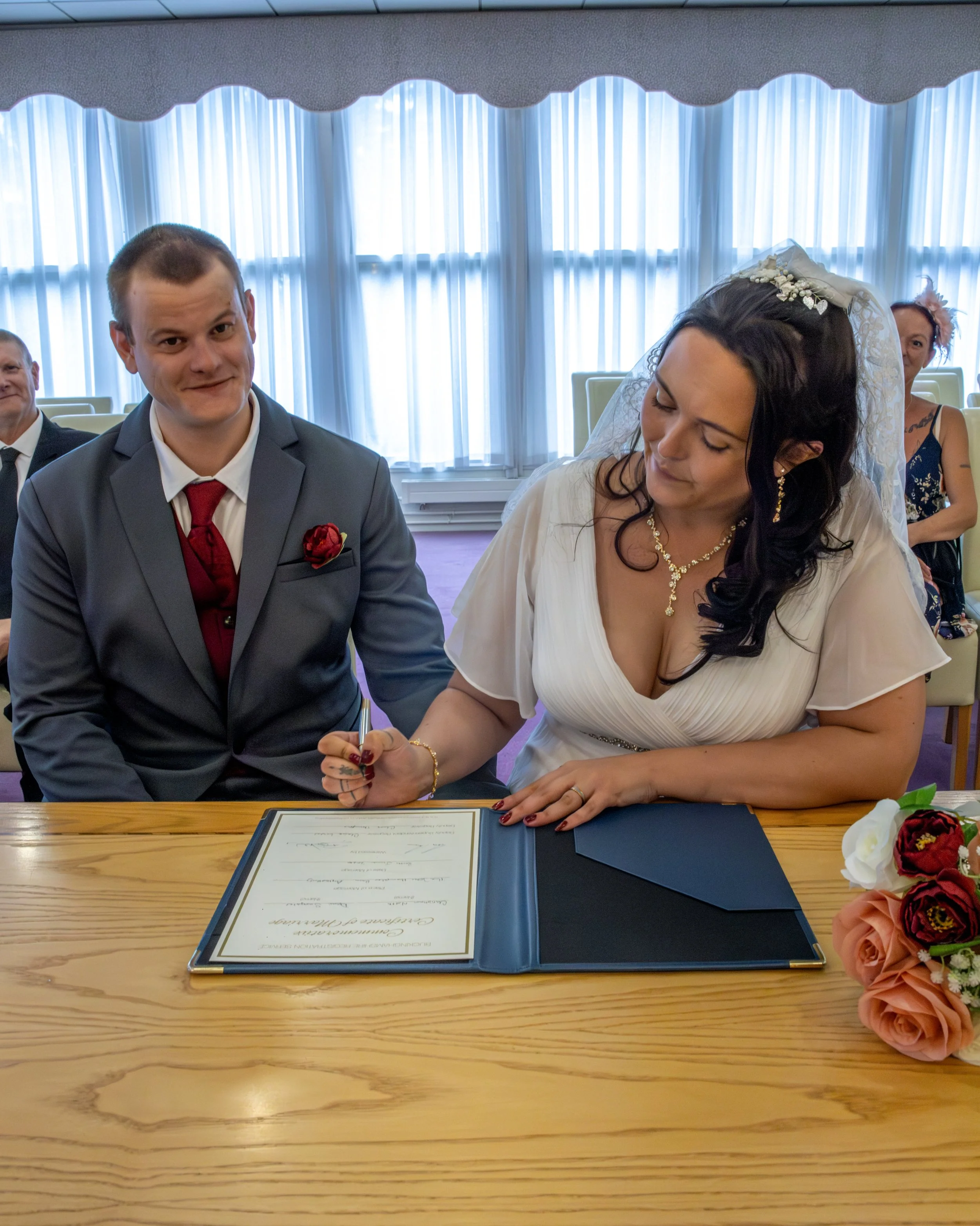 A couple is signing a marriage certificate at a wooden table during a wedding ceremony. The groom wears a gray suit with a red tie and boutonniere, while the bride wears a white dress with a pearl necklace and veil. Two women sit in the background, o