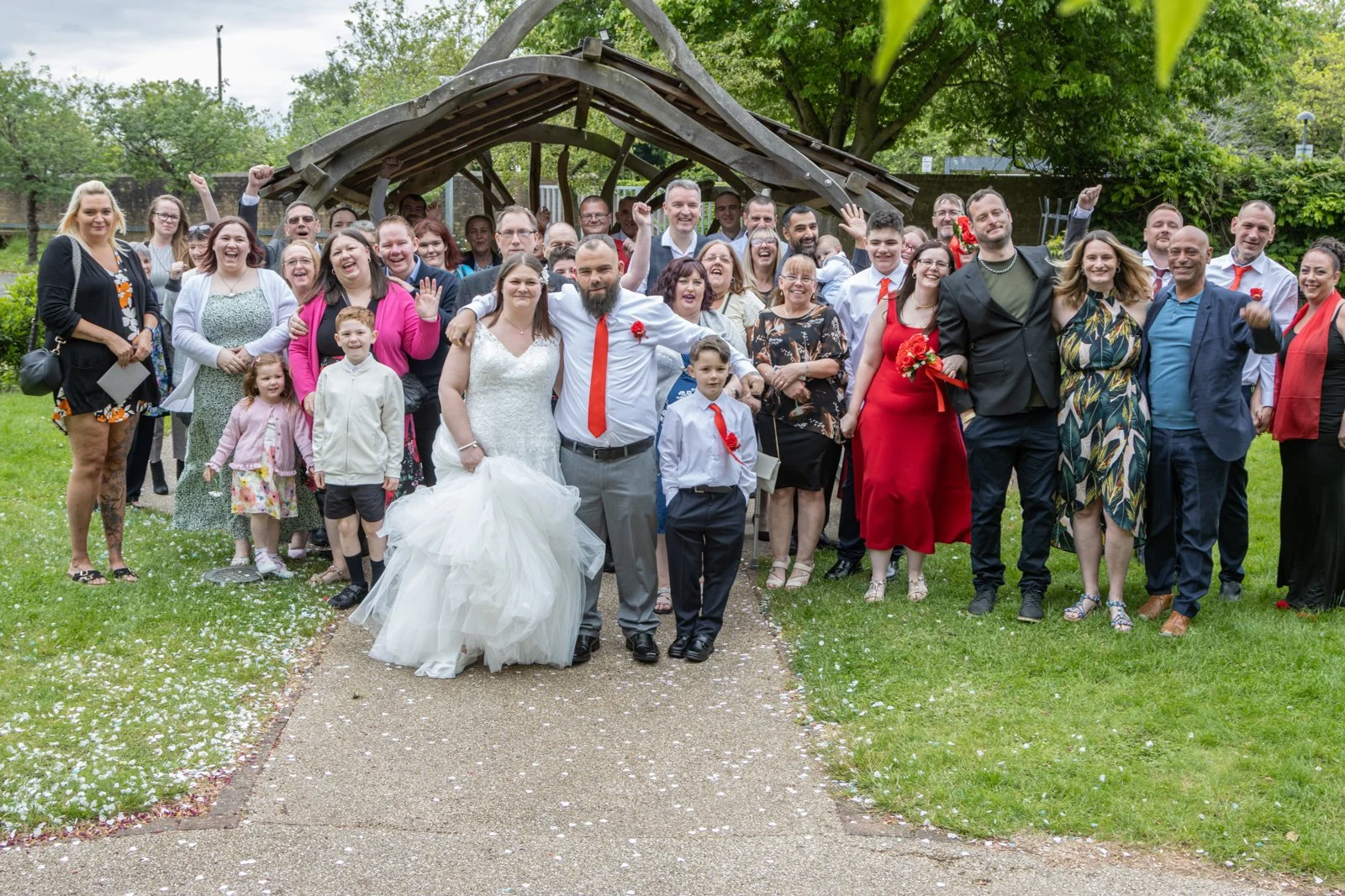A large group of people gathered outdoors for a celebration, including a bride and groom in wedding attire, with the bride in a white gown and the groom in a white shirt and red tie, standing in front. The group appears joyful, with some people smili