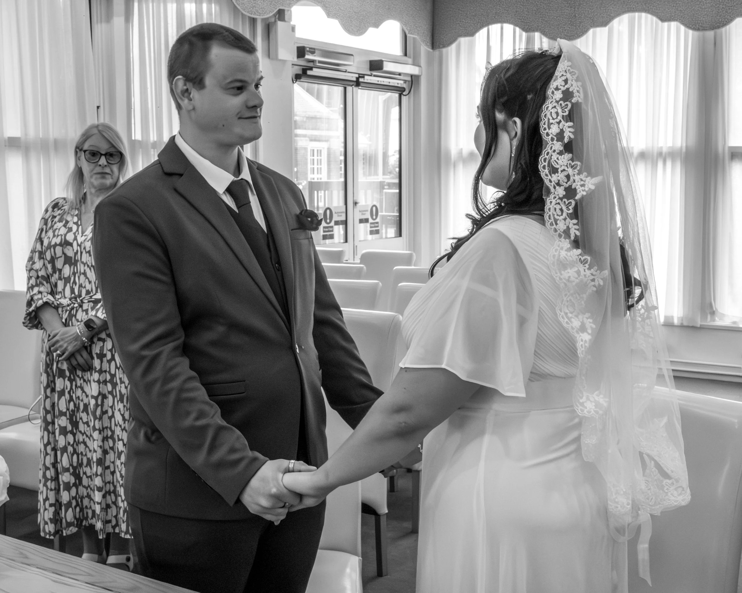 A wedding ceremony where a man in a suit is holding hands with a woman in a white dress and lace veil, facing each other, with a woman in glasses and patterned dress standing in the background inside a decorated room.
