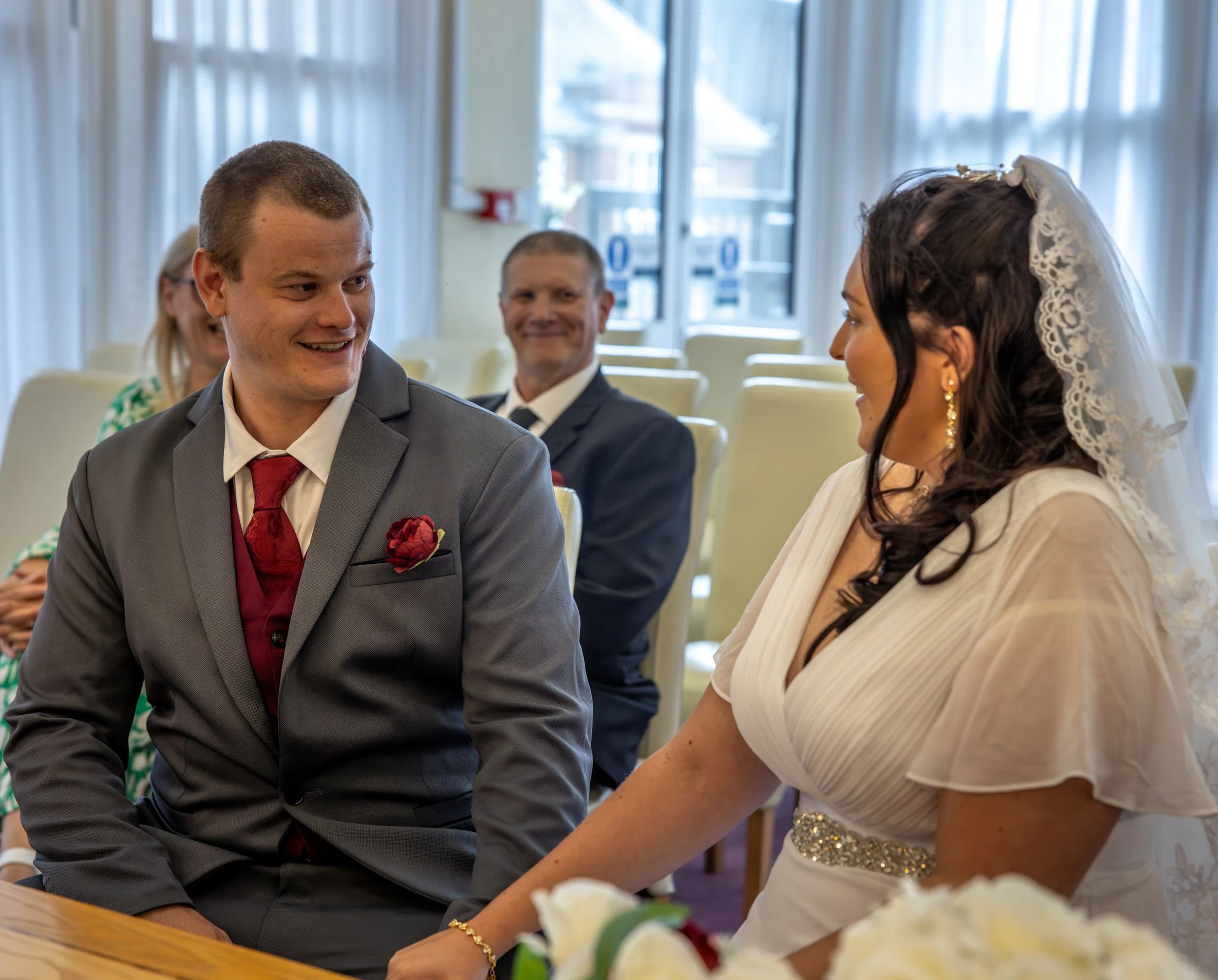 A bride & groom at a wedding ceremony, holding hands and looking at each other, with guests seated in the background.