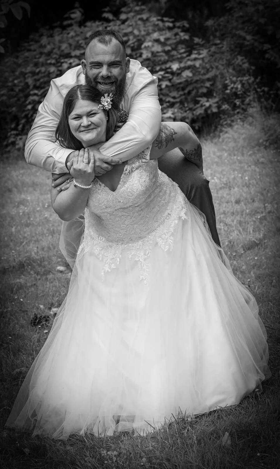 A joyful couple in wedding attire, smiling and embracing outdoors on a grassy area with trees in the background; woman in a lace wedding gown with a floral hair accessory, man in a white shirt with tattooed arms.