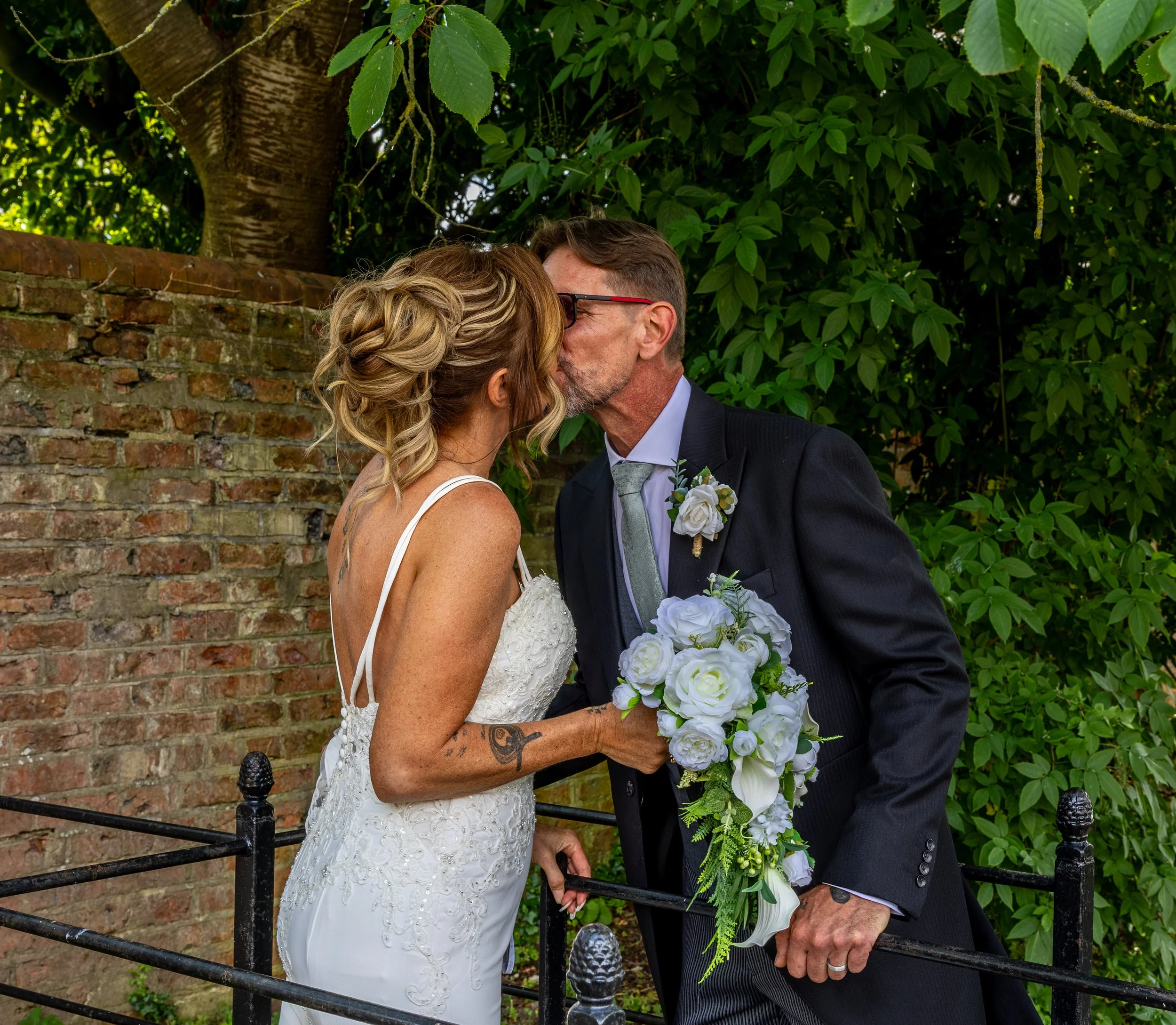 A bride and groom sharing a kiss outdoors during their wedding. The bride is wearing a white lace wedding dress and has blonde, styled hair. The groom is wearing a black suit with a green tie and glasses. They are holding hands, and the bride is hold