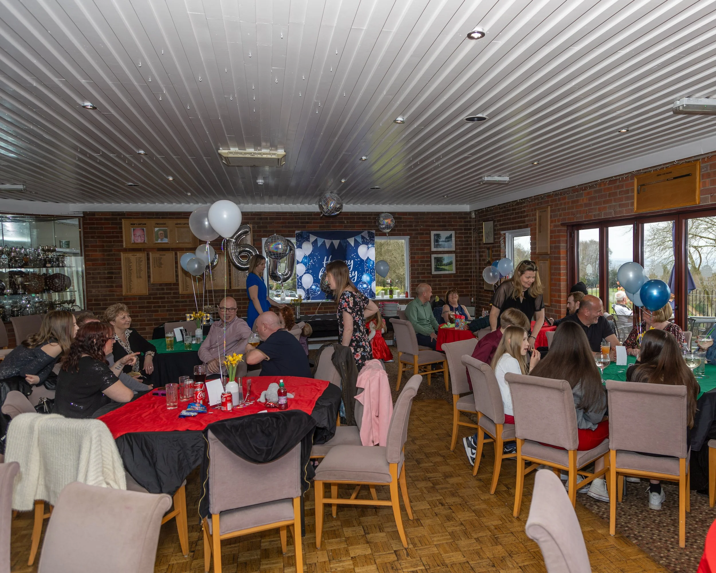 People celebrating a birthday in a decorated indoor hall with tables, balloons, and a banner that reads 'Happy Birthday'.
