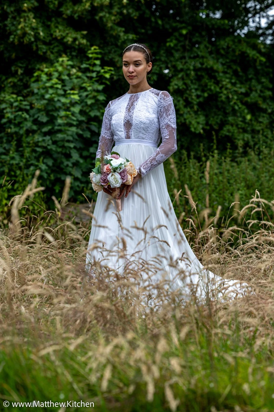 Bride in a white wedding dress with a lace top and long sleeves, holding a bouquet of pink and white roses, standing in a grassy field with green trees in the background.