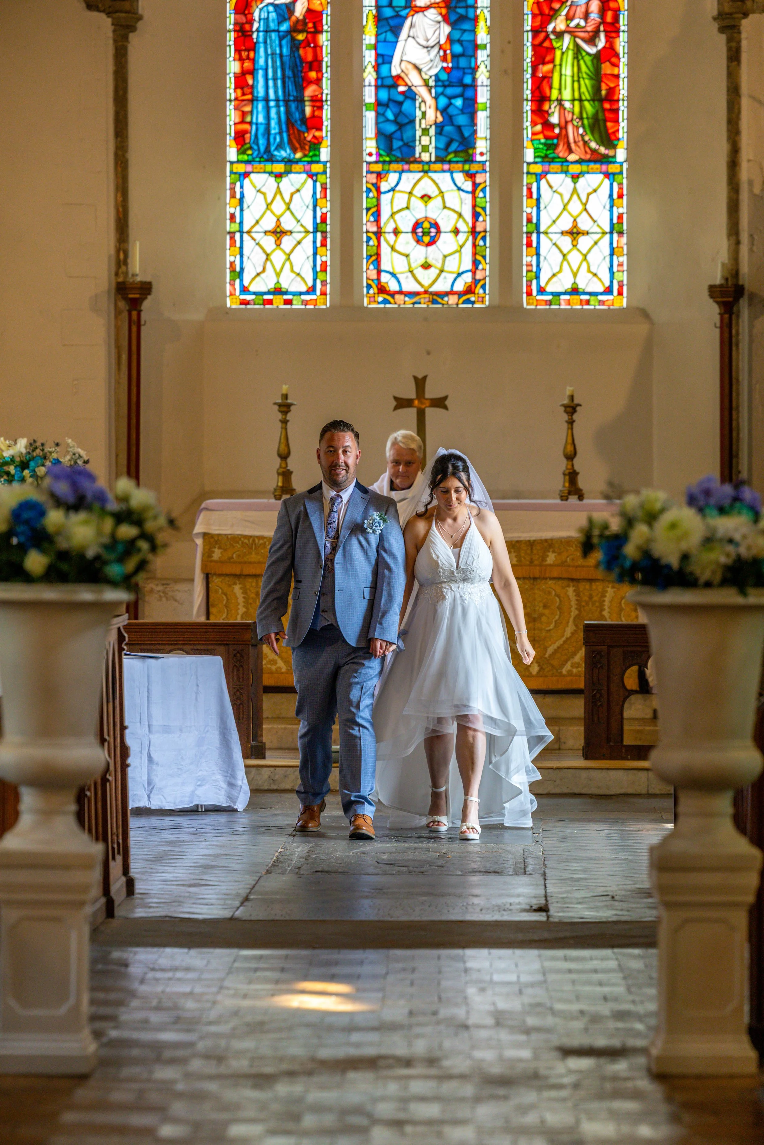A bride and groom walking down the aisle in a church with stained glass windows depicting religious figures, an altar, and floral arrangements.