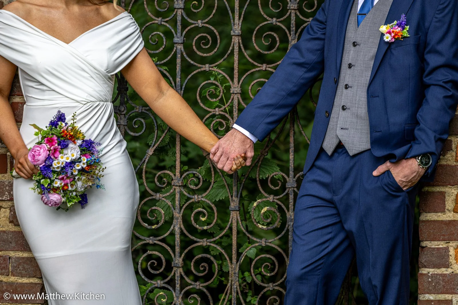 A couple holding hands, with the bride in a white dress holding a colorful bouquet, and the groom in a navy blue suit with a floral boutonniere. They are standing outdoors near a decorative metal fence.