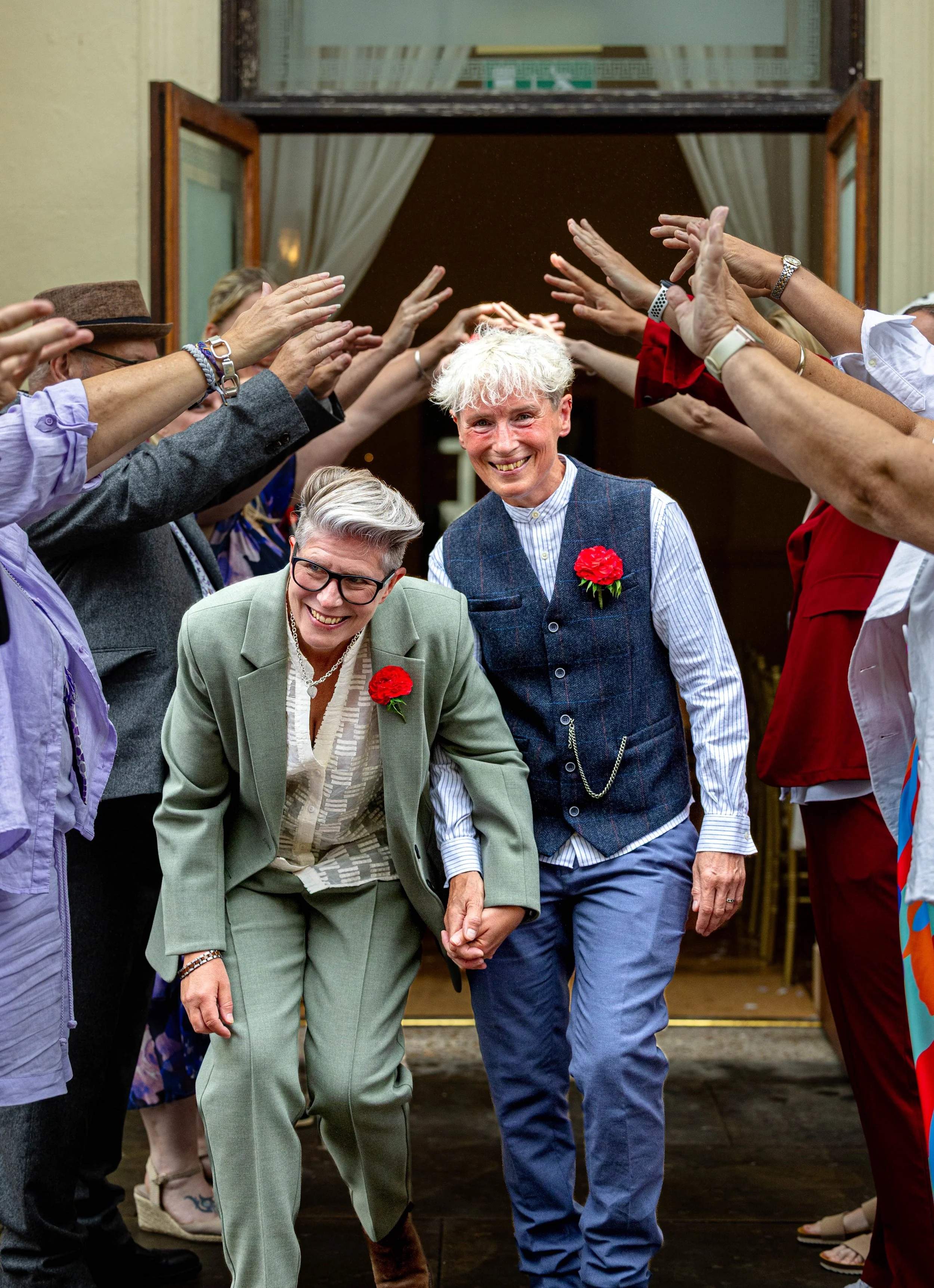 Two women holding hands and smiling, walking through an arch of raised hands during a celebration outside a building, with one woman wearing a grey suit and glasses, and the other wearing a dark vest over a striped shirt, both with red flowers pinned