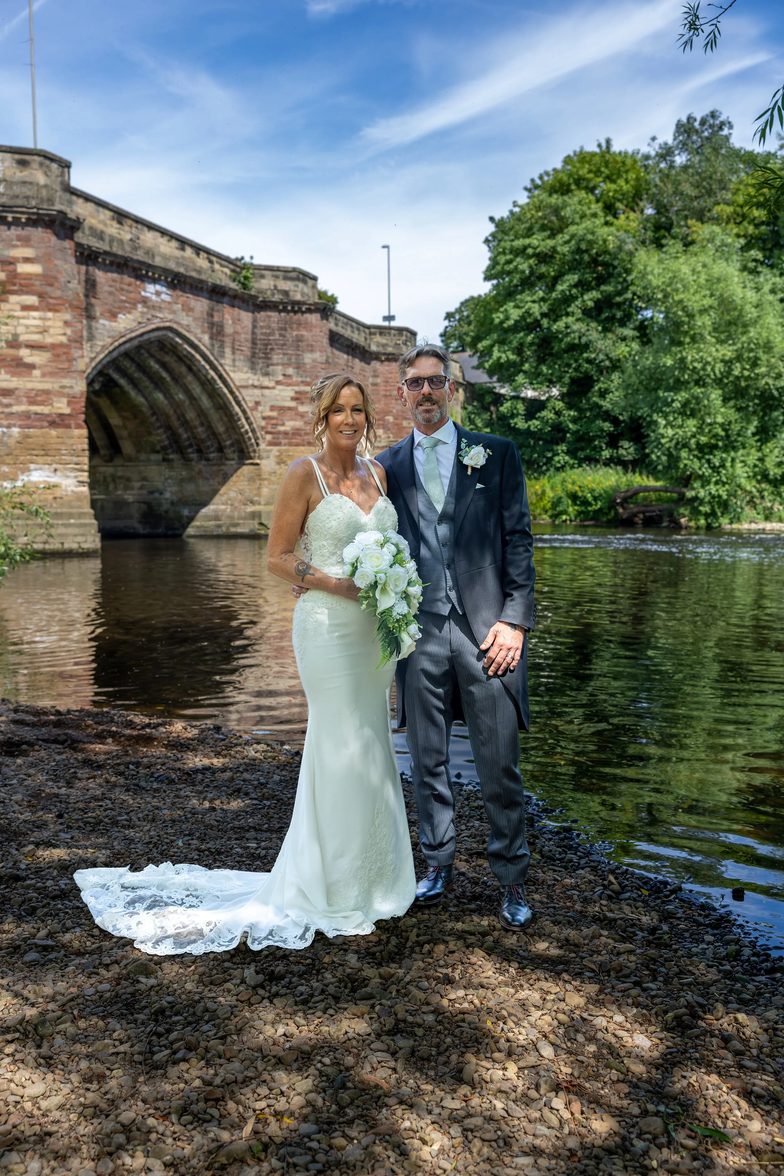 A bride and groom standing together on a rocky riverside, with a historic brick bridge and greenery in the background, under a partly cloudy sky.