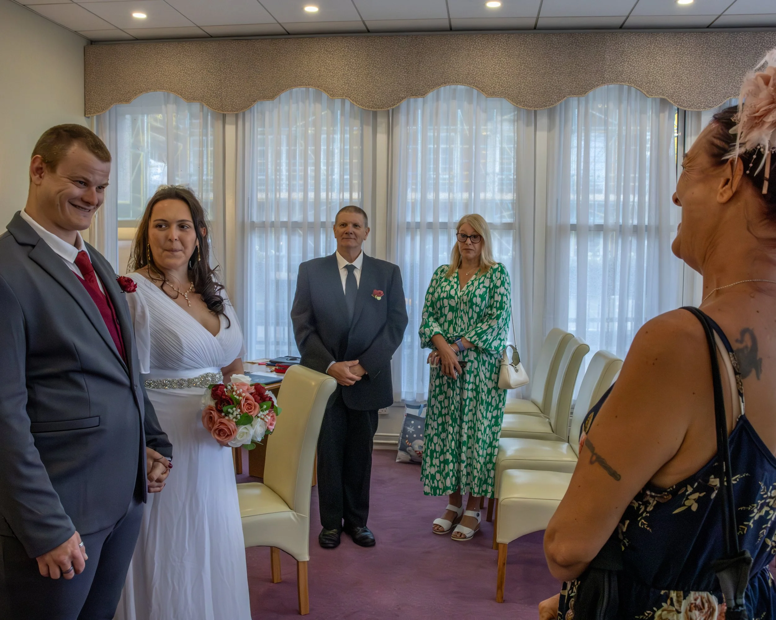 A wedding ceremony with a bride in a white dress holding a bouquet of flowers, standing next to a groom in a suit. Four other guests, two men and two women, are standing nearby inside a room with large windows and curtains.