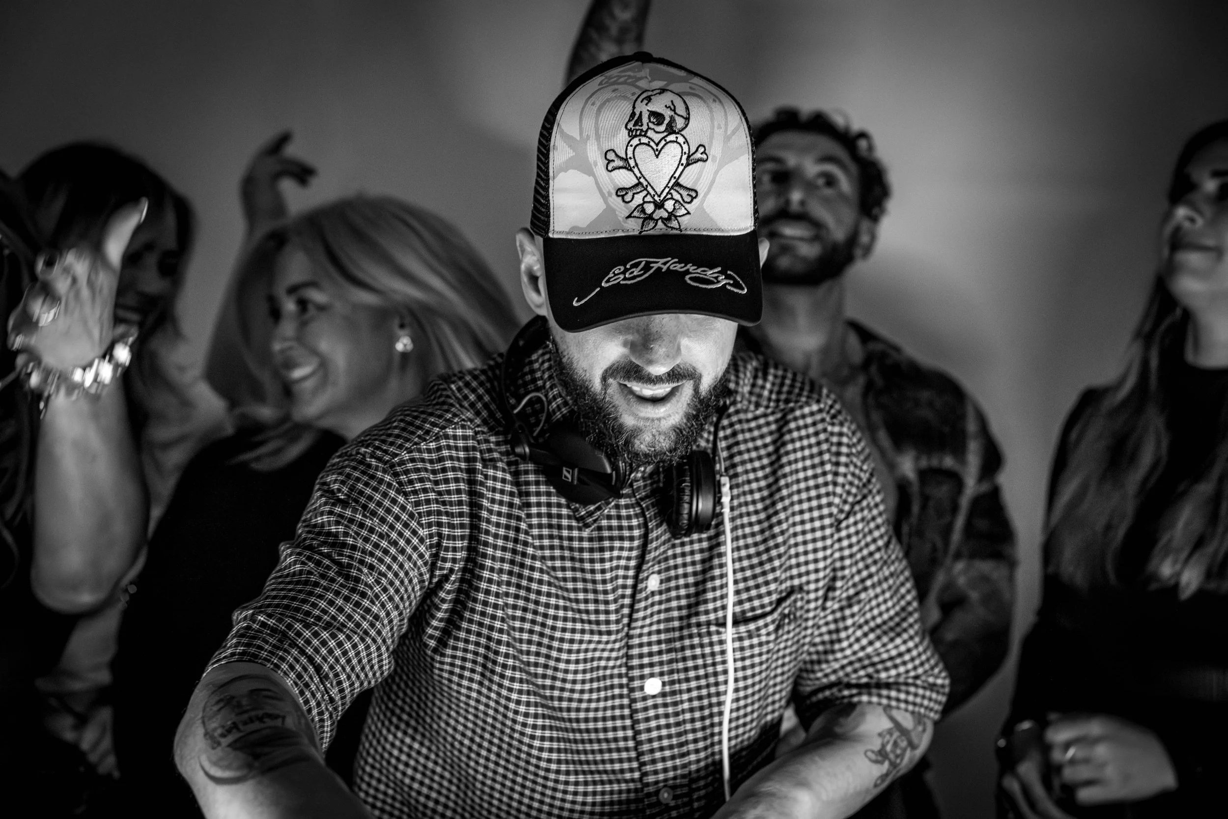 Black and white photo of a group of people, with a man in a checkered shirt and a cap with a heart and skull design, smiling and surrounded by women who are talking and laughing.